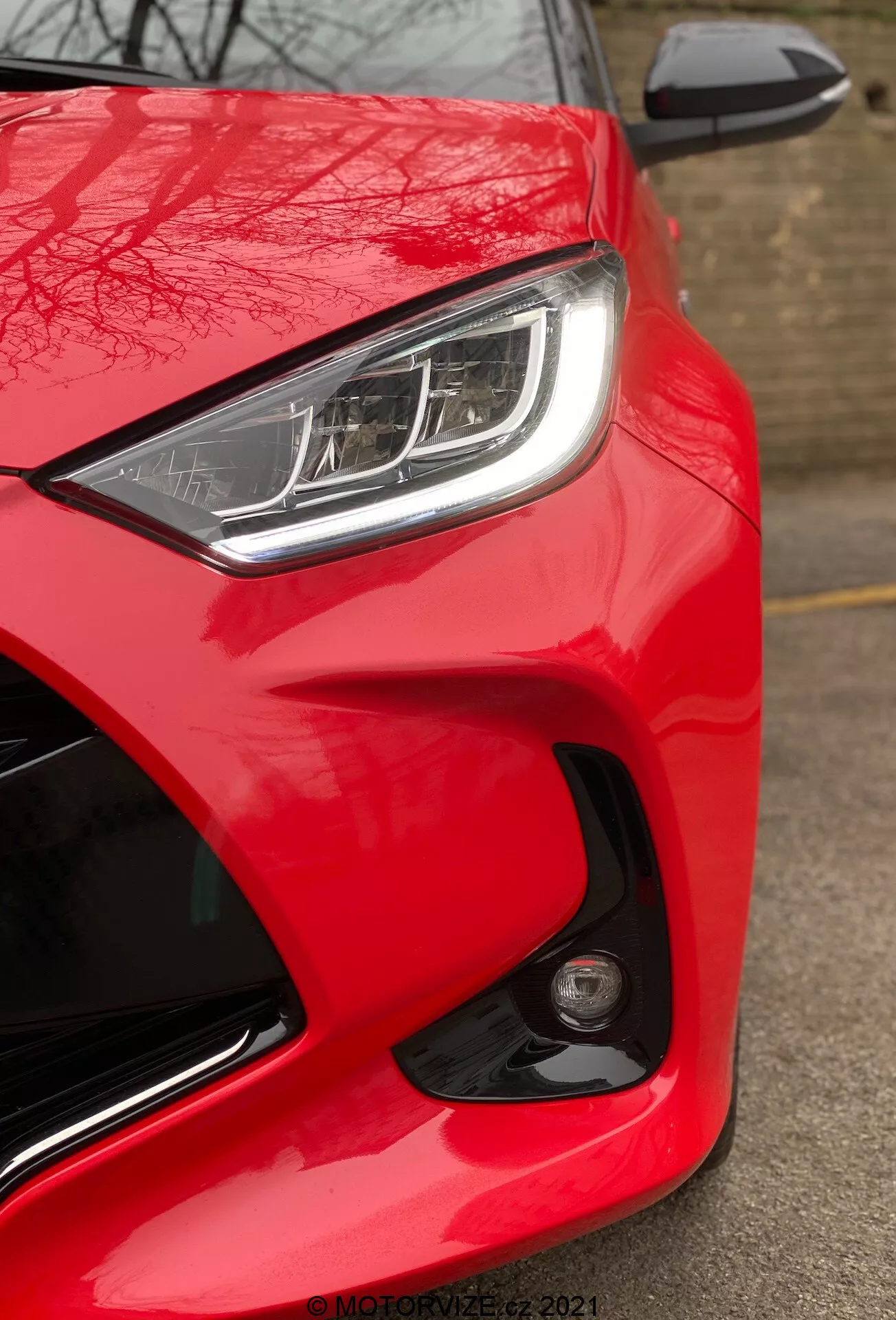 Od blizu prikaz Toyote Yaris 2020, osredotočen na sprednji konec vozniške strani z nizkega kota. Slika poudarja svetlo, sijoče rdečo barvo avtomobila, elegantni LED ali HID žaromet z značilnimi dnevnimi lučmi in delno vidno stransko ogledalo v barvi karoserije z vgrajeno signalno lučjo. Aerodinamični dizajn je poudarjen s izklesanimi linijami in krivuljami okoli žarometa in pokrova motorja. Okrogla meglenka ali senzor na sprednjem odbijaču nakazuje napredne varnostne funkcije. Pomanjkanje zameglitve gibanja nakazuje, da je avto stacionaren, desno spodaj pa je viden vodni žig 'MOTORZETZ. 2021'.