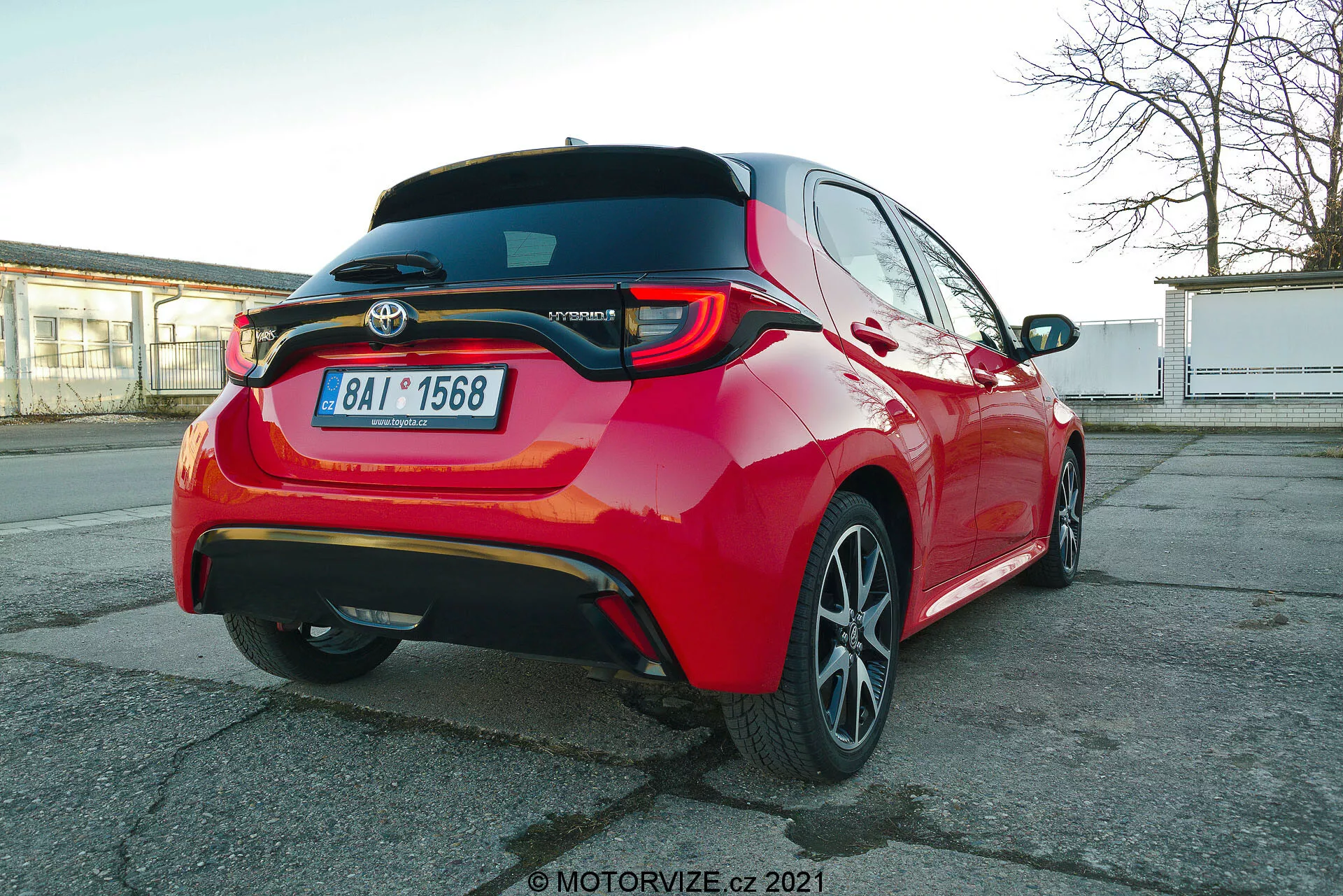 Vibranten rdeč osebni avtomobil Toyota Yaris 2020 fotografiran iz zadnjega tričetrtinskega kota, izpostavlja LED zadnje luči, zadnji spojler, embleme hibridnega modela, večkraka lita platišča in vidne funkcije za pomoč vozniku, kot so parkirni senzorji in zadnja kamera, zajete iz nekoliko nižje višine vozila.