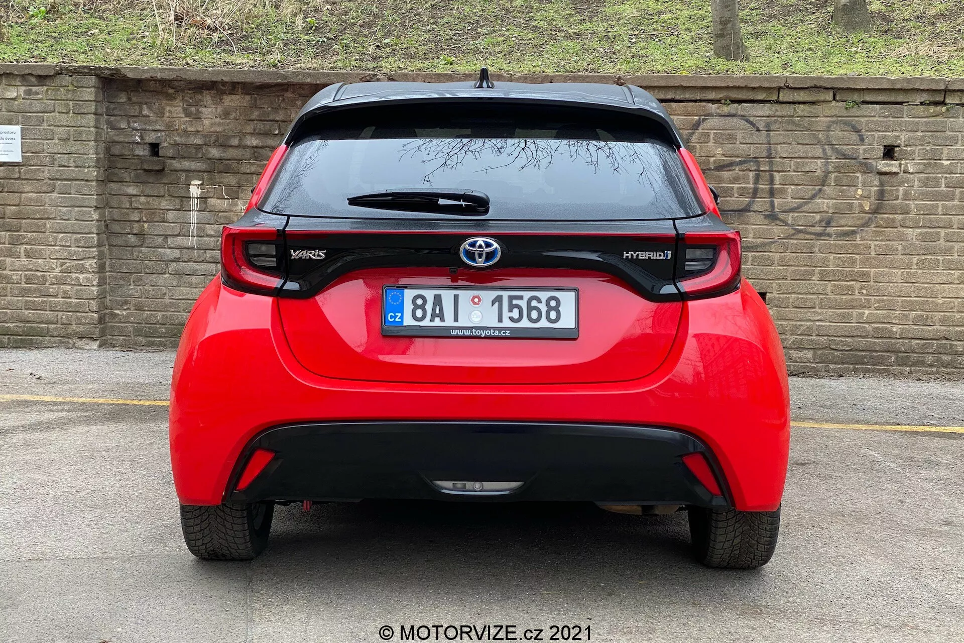Zadnji pogled na rdečega Toyota Yaris Hybrid iz leta 2020, osredotočen in posnet pod neposrednim kotom. Slika razkriva celotno širino vozila, prikazuje znak Toyota, imeni modelov 'Yaris' in 'Hybrid', spojler, strešno nameščeno anteno, visoko nameščeno zavorno luč in značilne zadnje luči. Vidno je tudi zadnje okno z brisalcem in spodnji del odbijača, ki verjetno vsebuje senzorje ali odsevnike. V ozadju je opečnata stena ali ograja in nekaj rastlinja, kar nakazuje nižje parkirano vozilo ali nižji kot kamere.