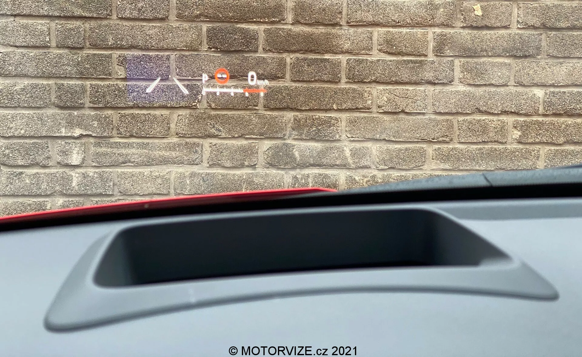 Pogled od blizu iz notranjosti Toyote Yaris 2020, ki kaže delni pogled s sedeža voznika nazaj. Poudarek je na zadnji polički z visoko nameščenim zavorno lučko in integriranim zadnjim spojlerjem. Slika prikazuje tudi del notranjih materialov, medtem ko opečna stena zunaj kaže, da je avto parkiran blizu stavbe.