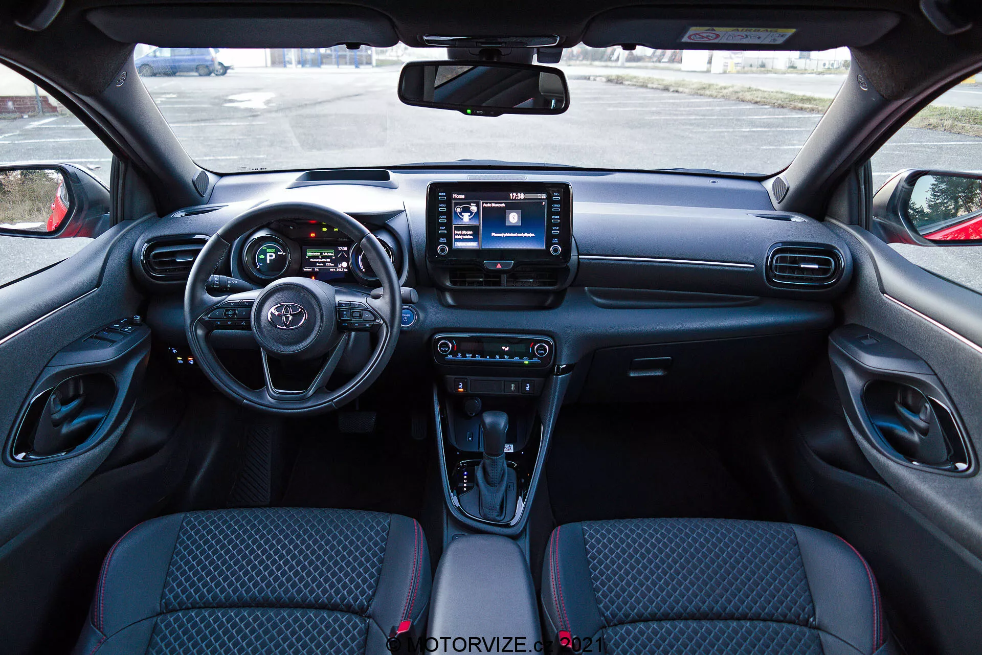 Pogled na notranjost Toyota Yaris 2020 iz perspektive voznikovega sedeža, z moderno armaturno ploščo s volanom, ki ima vgrajene kontrole, delno vidno instrumentno ploščo z digitalnimi zasloni in centralnim sistemom infotainmenta z zaslonom na dotik. Armaturna plošča vsebuje zračne reže in gumbe za nadzor klime, medtem ko ima sredinska konzola kontrole za načine vožnje, samodejni menjalnik z usnjenim oblazinjenjem, držala za pijačo in predal za shranjevanje. Sedeži iz blaga imajo vzorčast dizajn, notranji paneli vrat pa vključujejo kontrole za električne okna in nastavitve ogledal.