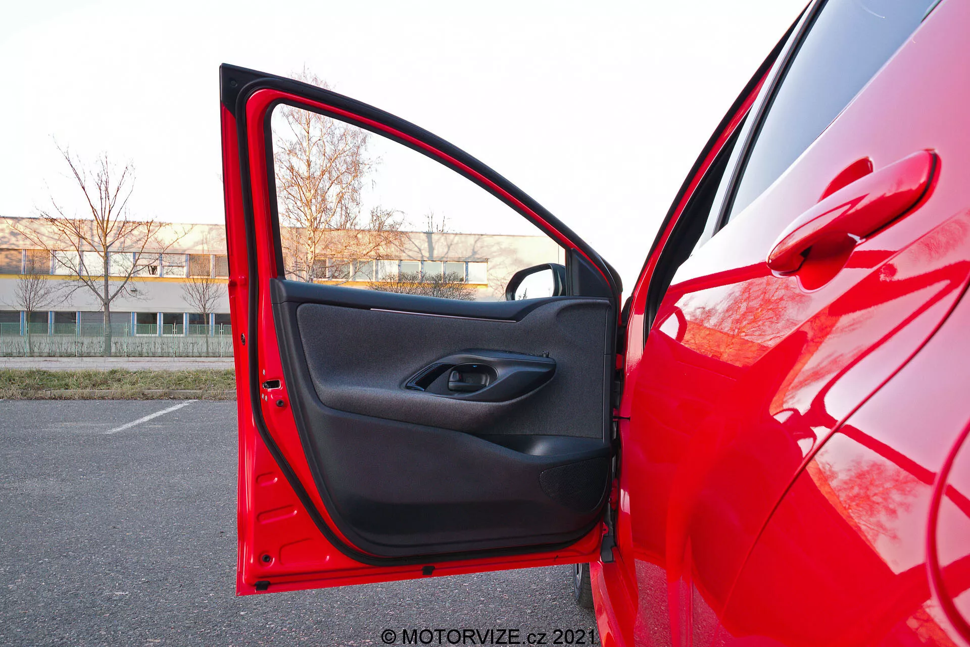 Bližnji pogled na rdečo Toyota Yaris 2020 s strani voznika z odprtimi sprednjimi vrati, posnet iz rahlo nizkega kota. Kamera je postavljena zunaj, blizu vrat, usmerjena proti notranjosti, prikazuje svetlo rdečo zunanjo barvo, ki odbija svetlobo, in črno notranjo oblogo z minimalističnim dizajnom. Vrata imajo gumb za električni nadzor oken znotraj vdolbinice ročaja in spodaj žep za shranjevanje.