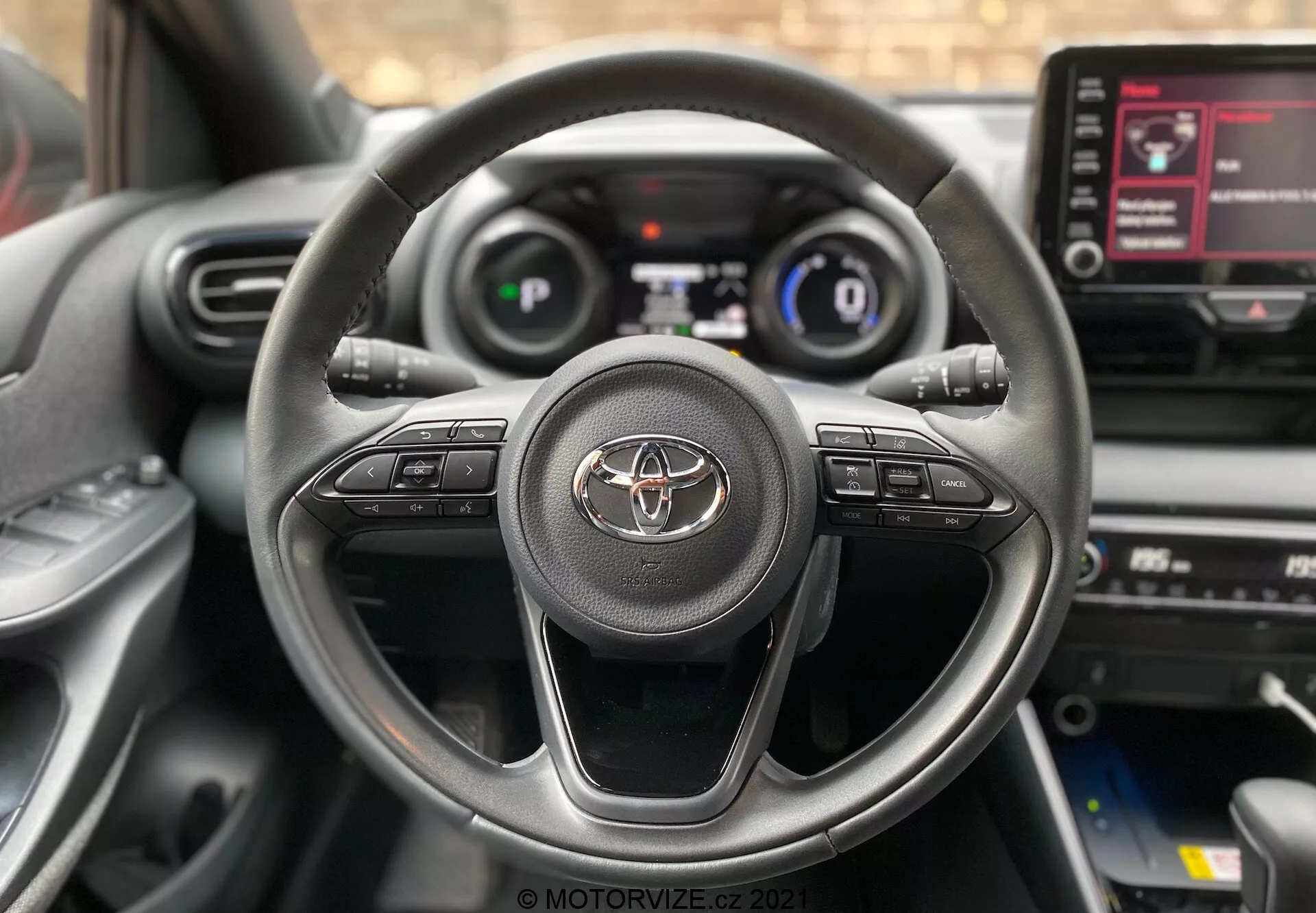 Priblžan pogled na notranjost Toyota Yaris 2020 iz voznikovega sedeža, s trikraktim volanom z logotipom Toyota in gumbi za nadzor avdio sistema, klicev prostoročne telefonije in tempomata. Delno je viden digitalni prikazovalnik z merilnikom hitrosti in osnovnimi informacijami o vožnji. Vidna je tudi sredinska konzola z zaslonom na dotik za informacijsko razvedrilni sistem z menijskimi možnostmi.