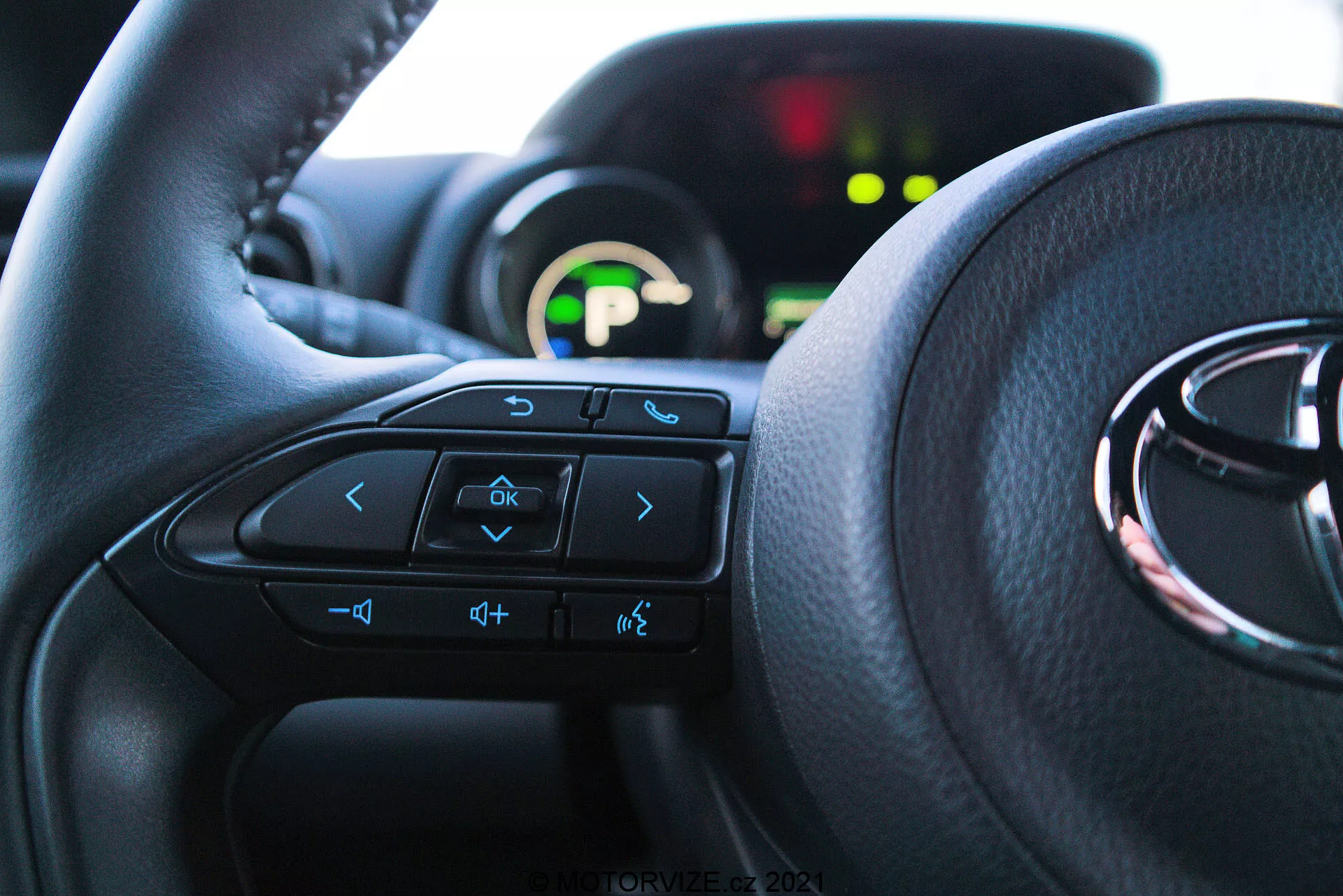 Bližnji notranji pogled na Toyota Yaris 2020 iz perspektive voznikovega sedeža, poudarjajoč volan z večfunkcijskimi kontrolami in instrumentno ploščo v ozadju. Volan dominira spodnji dve tretjini slike, vidni gumbi vključujejo puščice, glasovne ukaze, glasnost in gumbe za zabavni sistem. Delno vidna instrumentna plošča je osvetljena z zelenim digitalnim zaslonom in je izven fokusa zaradi plitve globinske ostrine. Naravna svetloba osvetljuje notranjost, kar poudarja udobje in uporabniku prijazen vmesnik.