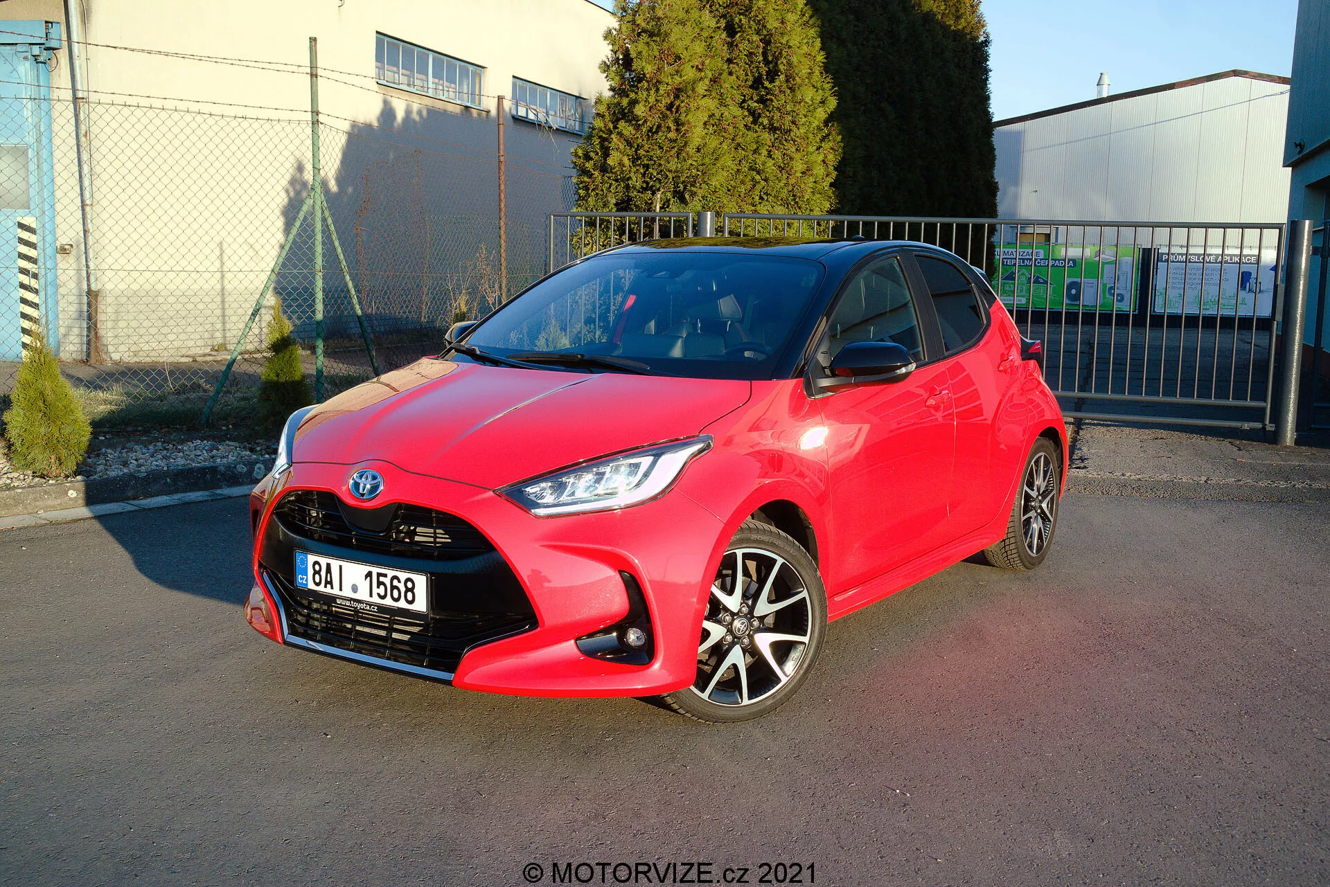 Slika svetlo rdečega hatchbacka Toyota Yaris iz leta 2020, parkirana pod kotom, ki prikazuje sprednji in stranski profil. Oblika žarometov je ostra in kotna, mreža ima vzorec satja s centralno postavljenim emblemom, sprednji odbijač pa ima vidne konture z meglenkami. Stranski profil razkriva dvobarvna aluminijasta platišča, barvno usklajene kljuke vrat in ogledala s čistimi linijami in obrisi vzdolž strani. Sijoča barva nakazuje novo ali dobro vzdrževano stanje. Ozadje vključuje jasno nebo, industrijske strukture in mehke sence, kar kaže na zunanje okolje, verjetno parkirišče ob komercialnem ali industrijskem objektu.