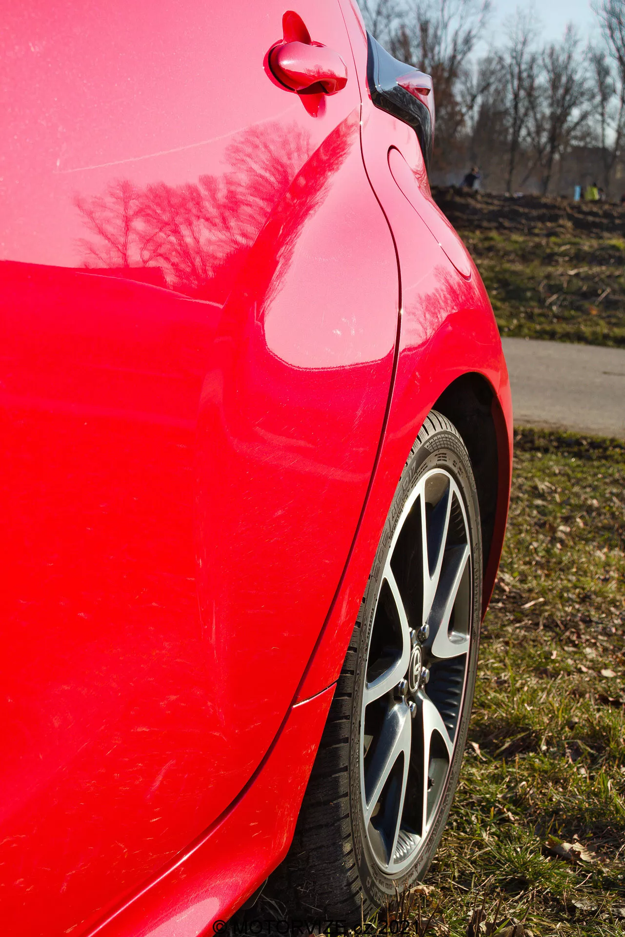 Bližnji bočni pogled na zadnji četrt panel rdečega Toyota Yaris 2020, posnet z rahlo navzgor usmerjenim kotom, poudarja svetlo rdečo barvo, večkratni prečno platišče, vzorec tekalne plasti pnevmatike, čist ukrivljenost zadnje četrtine panela in odbijača, bleščeč odsev barve z drevesi in jasnim modrim nebom ter vidno pokrov za gorivo in detajl okovja.
