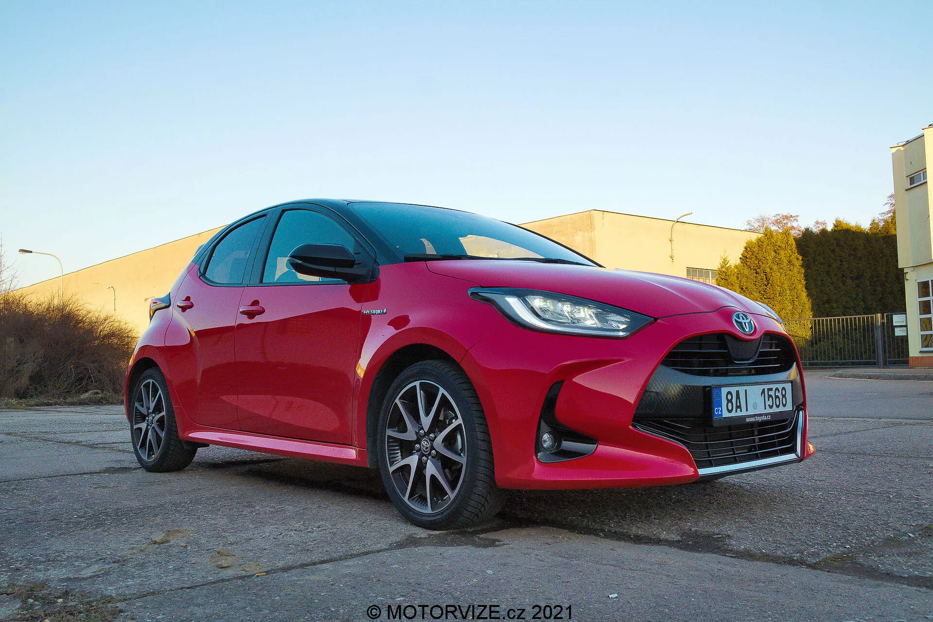 Nizkokotni pogled spredaj v treh četrtinah na rdeč Toyota Yaris 2020 hatchback, ki prikazuje ostre in kotne LED ali HID žaromete, izrazito mrežo s središčnim emblemom, večkrake aluminijaste platišča, integrirane smernike v stranskih ogledalih, čiste odbojne površine in možen zmogljiv zaviralni sistem. Posneto na toplo osvetljenem industrijskem parkirišču z vodnim žigom motorvize.cz 2021.