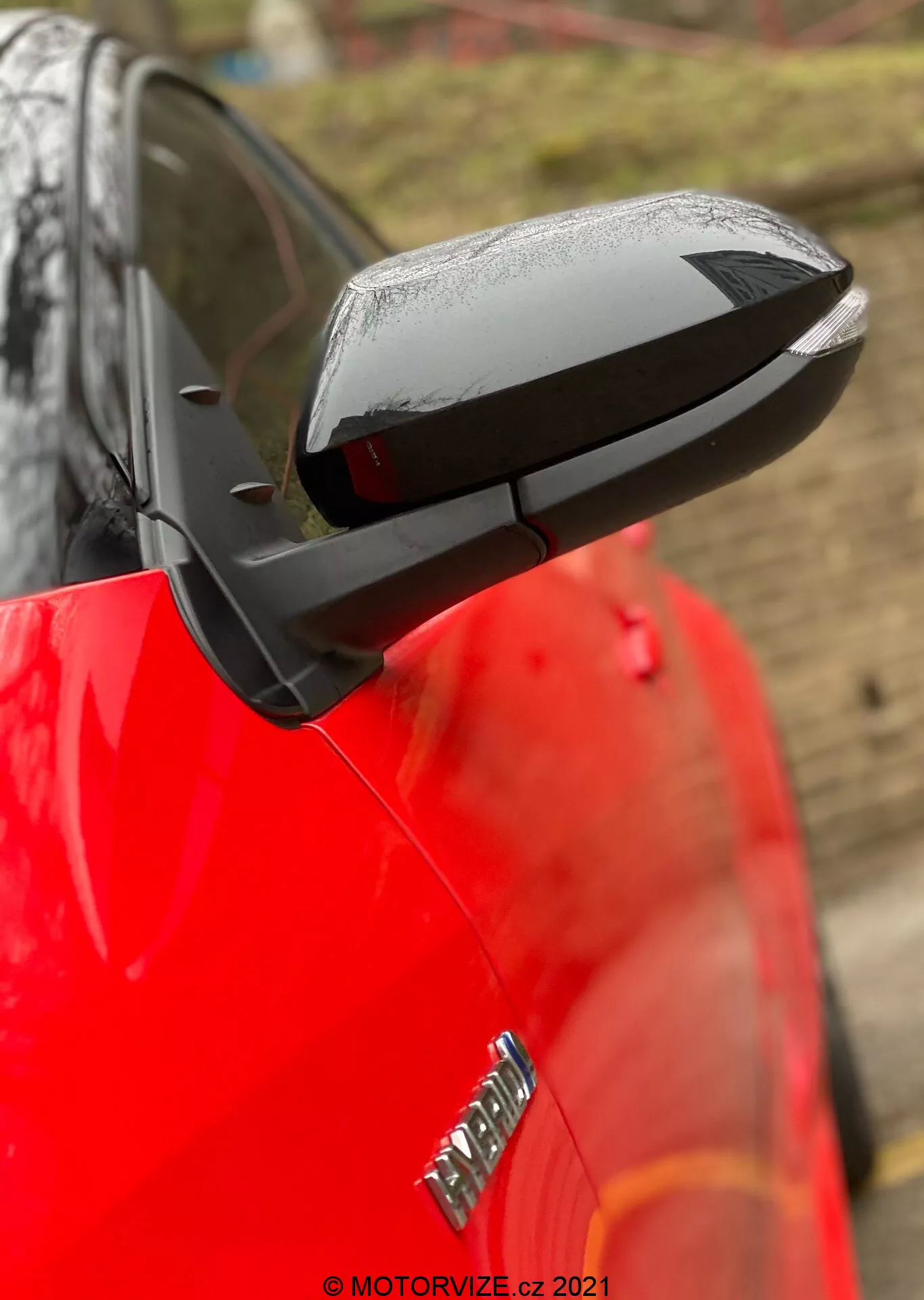 Pogled od blizu na raztegnjeno stransko ogledalo Toyote Yaris 2020 z elegantnim, aerodinamičnim črnim ohišjem, ki kontrastira z bleščečim rdečim karoserijo avtomobila. Ogledalo poudarja tehnološke funkcije, kot so možna električna nastavitev, gretje in vgrajeni smerniki, čeprav posebnosti niso potrjene. Kapljice vode kažejo na nedavni dež ali pranje avtomobila, medtem ko zamegljeno ozadje in plitka globina polja osredotočata pozornost na ogledalo. Del značke na sprednjem blatniku je viden, vendar je zamegljen.