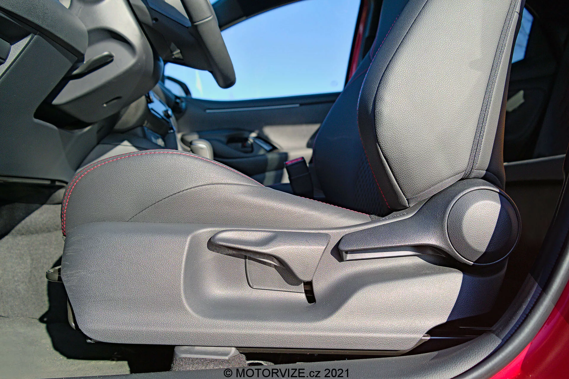 Notranji podrobni posnetek Toyote Yaris 2020, osredotočen na voznikova vrata in sedež, poudarja dvobarvno oblazinjenje, dekorativne šive, oblikovan dizajn za ergonomsko podporo, ročno nastavljive stranske opore in izrazito ročno ročico za nastavitev sedeža. Delno je viden okvir vrat, kar nakazuje, da so vrata odprta.