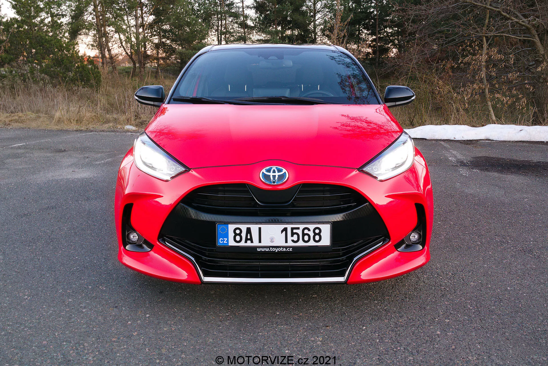 Slika prikazuje pogled od spredaj na rdečo Toyoto Yaris 2020, posneta je pod rahlim kotom, s poudarkom na sprednji maski, žarometih in pokrovu motorja, s popolnoma vidno registrsko tablico. Avto ima značilne oblikovalske elemente, vključno s Toyota emblemom sredinsko nameščenim na rešetki, ostrimi kotnimi žarometi, ki se zožujejo proti rešetki, in kompleksnimi krivinami na pokrovu motorja, kar prikazuje njegov aerodinamični in športni dizajn. Fotografija, posneta čez dan pred mrakom, nakazuje, da je avto parkiran na ravni površini v okolju, ki je lahko pozna jesen ali zgodnja pomlad.