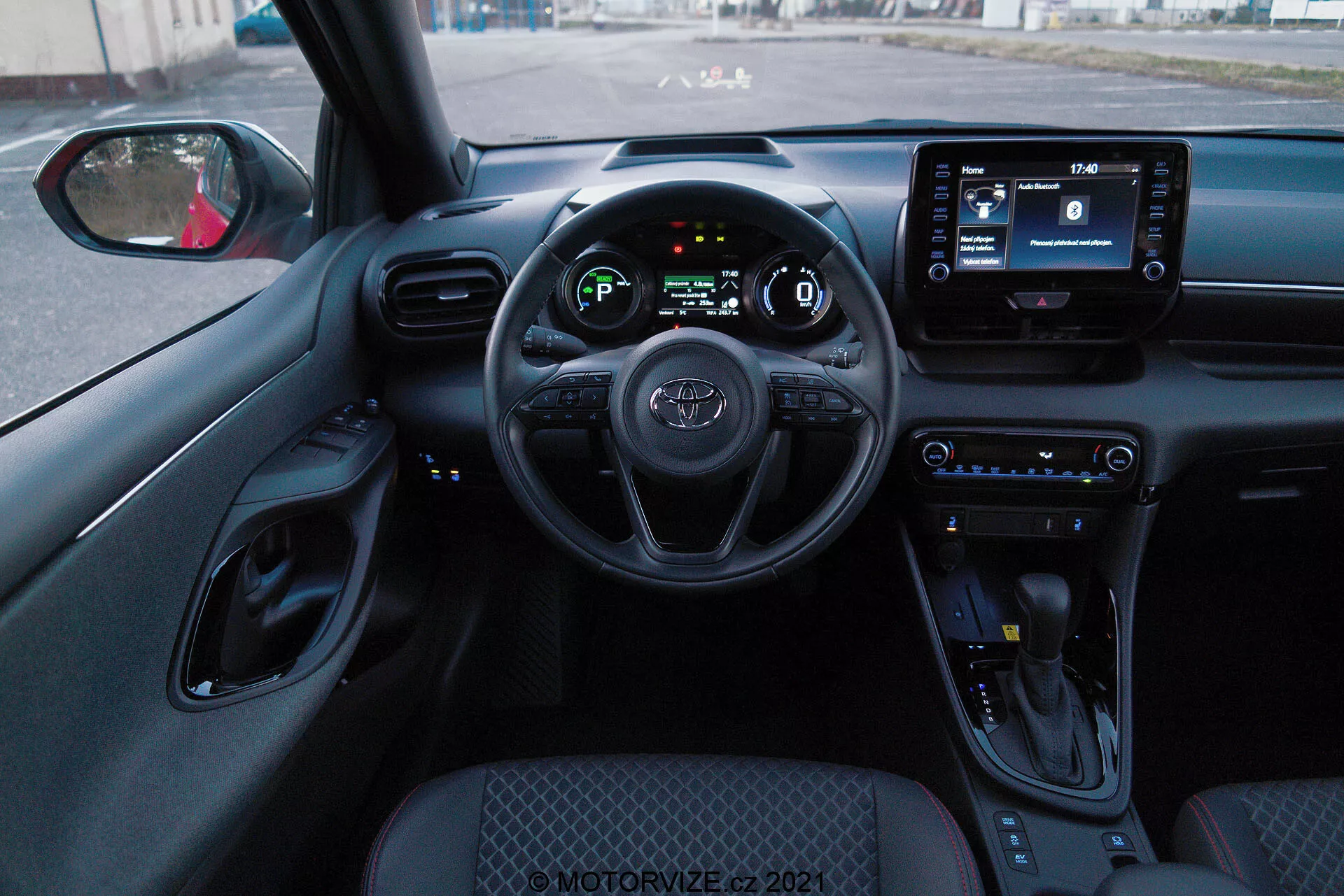 Perspektiva notranjosti s sedeža voznika v Toyoti Yaris 2020, ki prikazuje volan z nameščenimi kontrolami, digitalno instrumentno ploščo in infotainment zaslon. Notranjost vključuje gumbe za upravljanje klime, ročico menjalnika ter pretežno črno barvno shemo s teksturiranimi površinami in kromiranimi poudarki.
