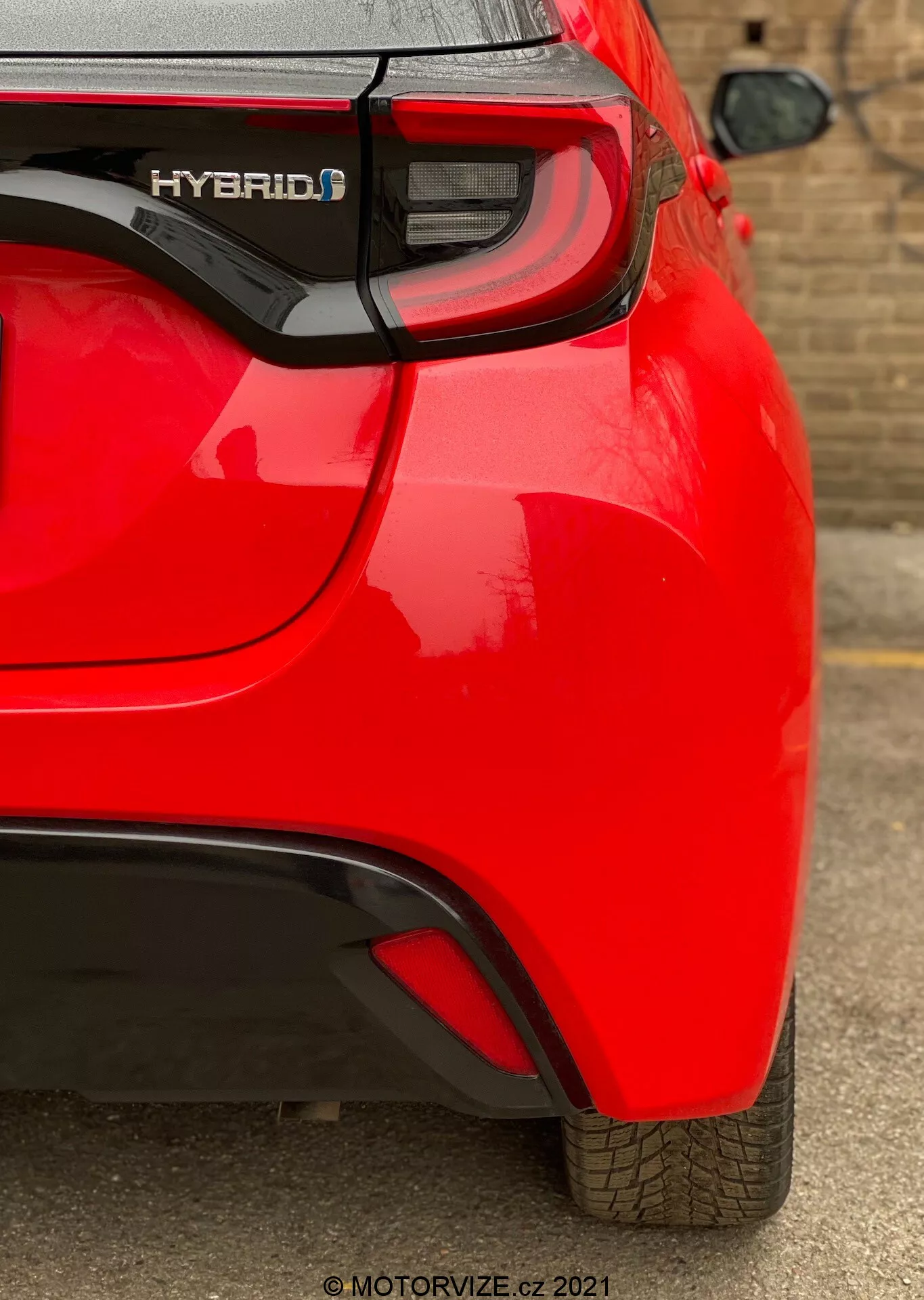 Bližnji, kotni zadnji pogled na rdeči hibridni avto Toyota Yaris 2020, s poudarkom na desnem zadnjem kvadrantu. Zajete so lastnosti, kot so oznaka 'HYBRID', sodobni LED zadnji svetlobni sklop, subtilni aerodinamični pregib na zadnjem odbijaču in viden parkirni senzor. Odsevna, sijoča barva in dobro ohranjen vzorec tekalne plasti nakazujeta nov ali dobro vzdrževan avto.