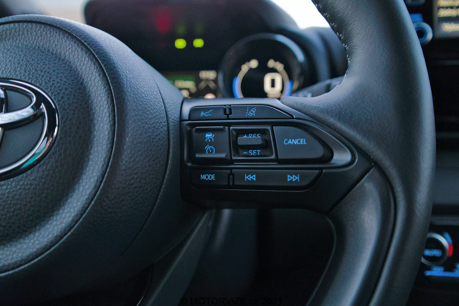 Posnetek notranjosti Toyota Yaris 2020 iz voznikovega sedeža, usmerjen na levo stran multifunkcijskega volana. Taktilni gumbi, vključno z ikono varnostne funkcije, tempomatom z oznako 'SET', gumbom za preklic, 'MODE' za multimedije in gumbi za glasnost '+', '-', so jasno vidni. Vozilo je v načinu 'P' parkiranja, pri čemer so instrumentna plošča in elementi v ozadju rahlo zamegljeni.