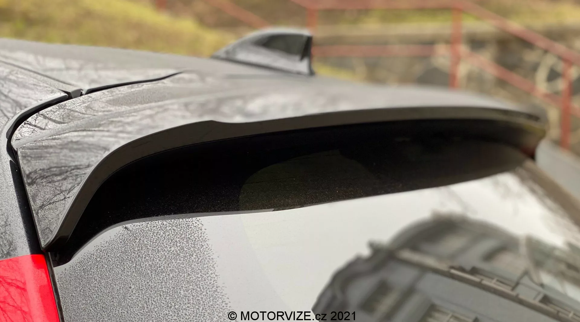 Bližnji posnetek zadnjega spojlerja Toyote Yaris 2020 s črno barvo, ki kontrastira z metalnim ohišjem. Slika ujame kapljice vode na spojlerju, kar kaže na mokre pogoje, in stranski profil, ki razkriva rahlo dvignjeno zasnovo za boljšo aerodinamiko. Zabrisano zunanje ozadje dodaja kontekst k premium finišu avtomobila, viden LED zadnji luč pa nakazuje sodobne funkcije.