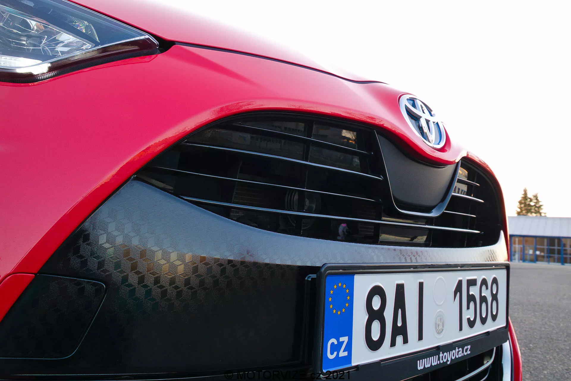 Posnetek od blizu rdečega Toyota Yaris 2020, osredotočen na levo sprednjo stran, prikazuje pokrov motorja, sodobno žaromet s potencialno LED ali HID tehnologijo, rešetko z znakom Toyota in vzorcem satovja, sijočo rdečo barvo ter delno vidni sprednji odbijač s spodnjim vstopom za zrak. Evropska registrska tablica označuje registracijo na Češkem.