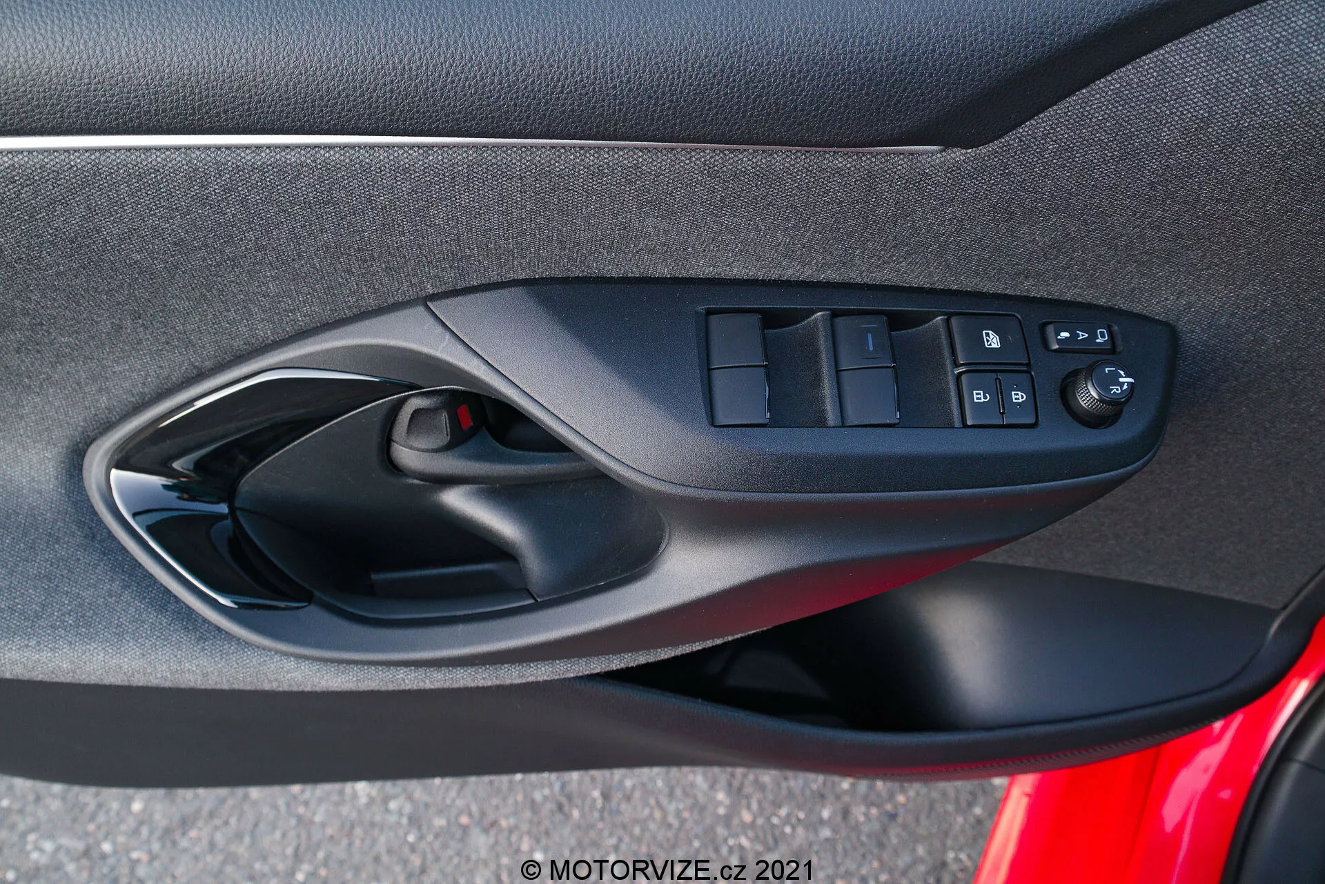 Notranji vratni panel Toyote Yaris 2020, osredotočen na ergonomsko oblikovane kontrole voznikovih vrat, vključno s stikali za električna okna, gumbom za zaklepanje vrat in električno nastavljivimi kontrolami ogledal, posnet iz perspektive potnika v vozilu. Fotografija poudarja mešanico mehkih tkaninskih ali kompozitnih materialov, črno barvno shemo in vidno mrežico zvočnika kot del avtomobilskega avdio sistema.