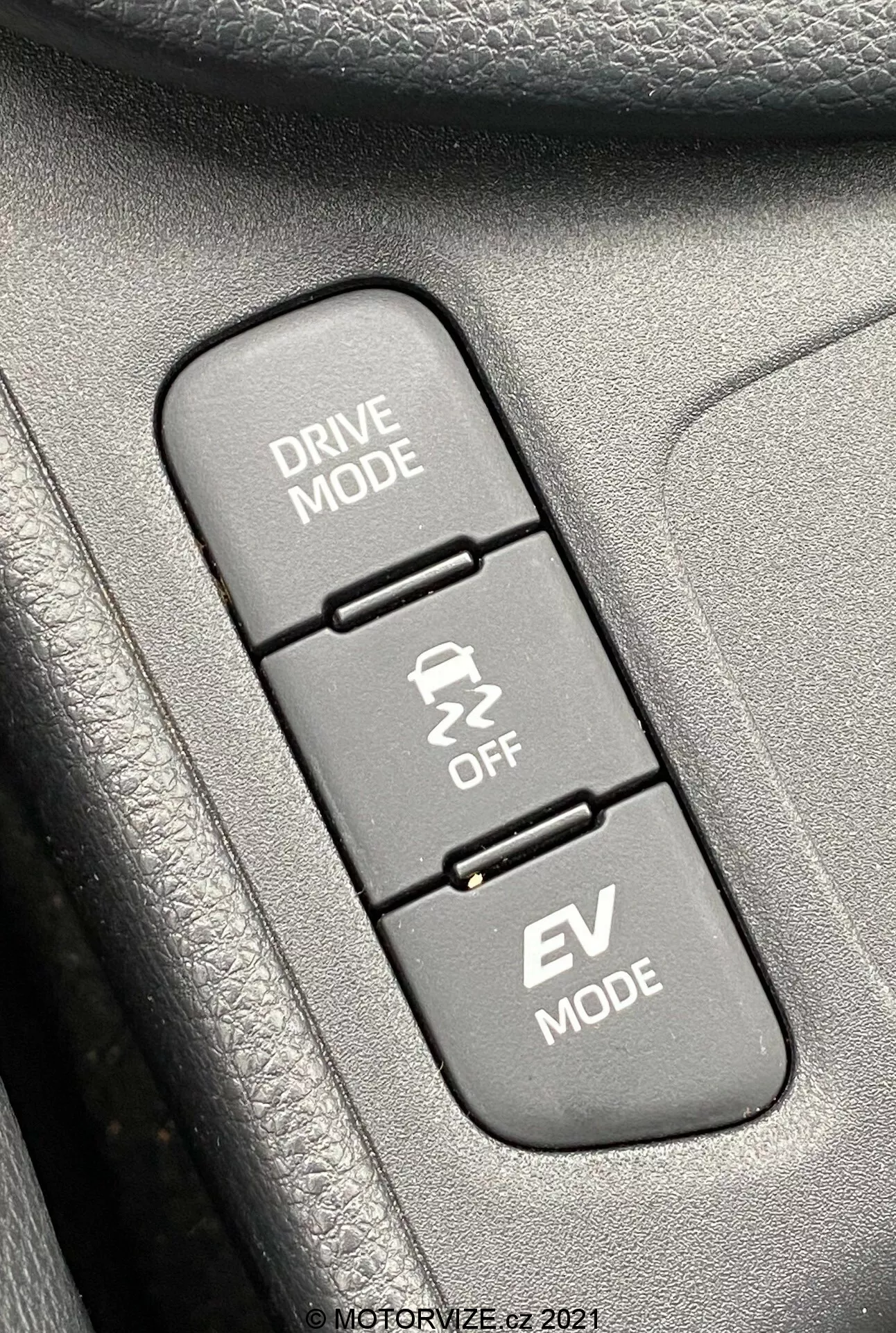 Povečan posnetek notranjosti Toyote Yaris 2020, osredotočen na tri navpično poravnane nadzorne gumbe, povezane z načini vožnje. Od zgoraj navzdol: 'DRIVE MODE' z ikono vijugaste ceste, 'Traction Control OFF' z ikono drsečega avtomobila in 'EV MODE', ki označuje funkcijo električnega vozila. Gumbi so nameščeni na teksturirani površini, verjetno del vratne plošče ali sredinske konzole. Slika je ostro osredotočena na gumbe, z okoliškimi teksturami rahlo zamegljenimi, enakomerno osvetljena z dnevno svetlobo za jasno vidljivost.