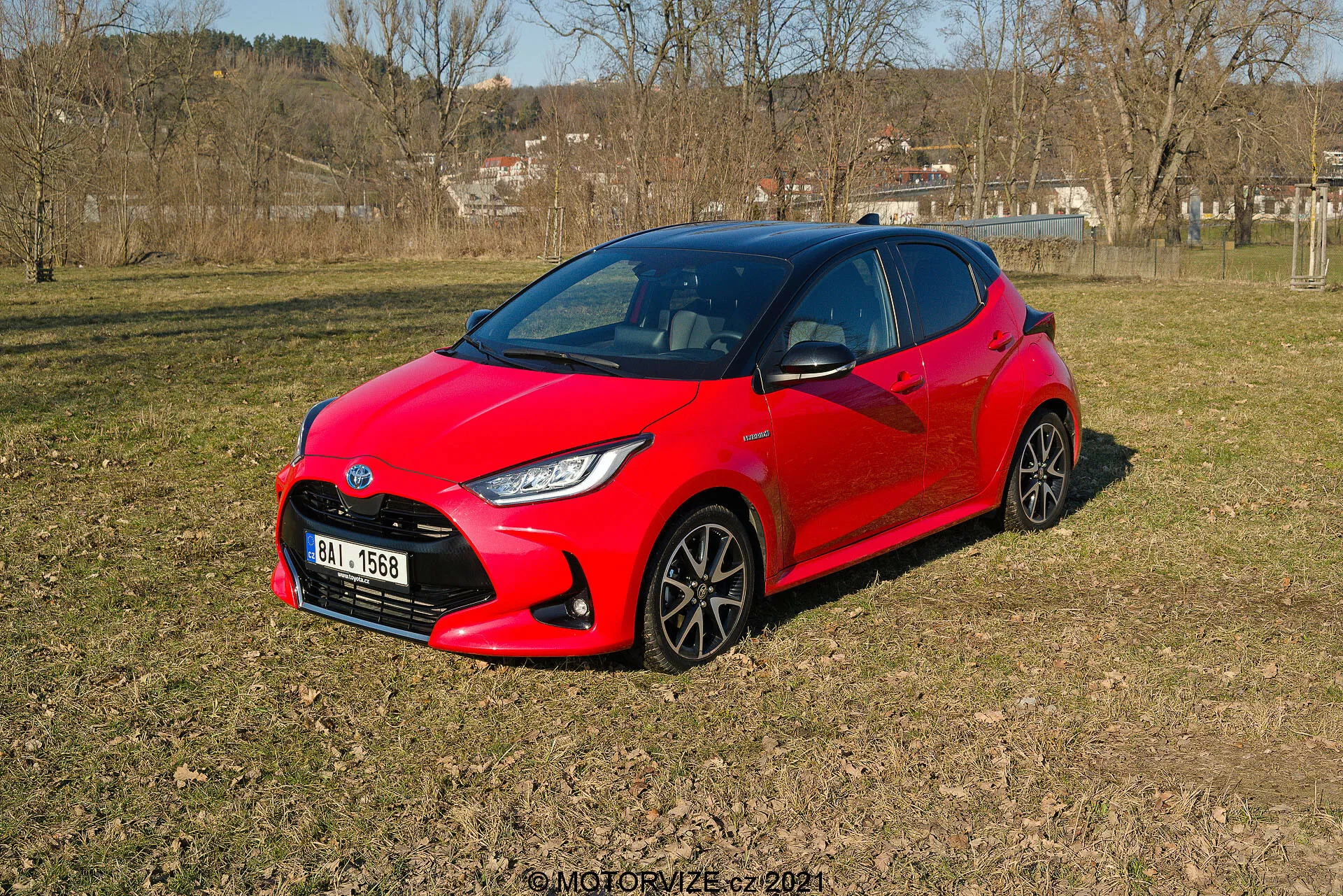 Slika prikazuje živahno rdeč Toyota Yaris 2020, parkiran na neenakomernem travnatem terenu v polpodeželskem okolju, gledano iz tridelne perspektive. Moderno, aerodinamično oblikovanje avtomobila vsebuje ostre kote okoli žarometov in kompaktno, športno podobo. Vidna so temna večkraka kolesa in stilizirani LED žarometi. Stranska okna in prednje vetrobransko steklo nudijo temen pogled v notranjost, kar nakazuje na možne napredne avtomobilske sisteme.