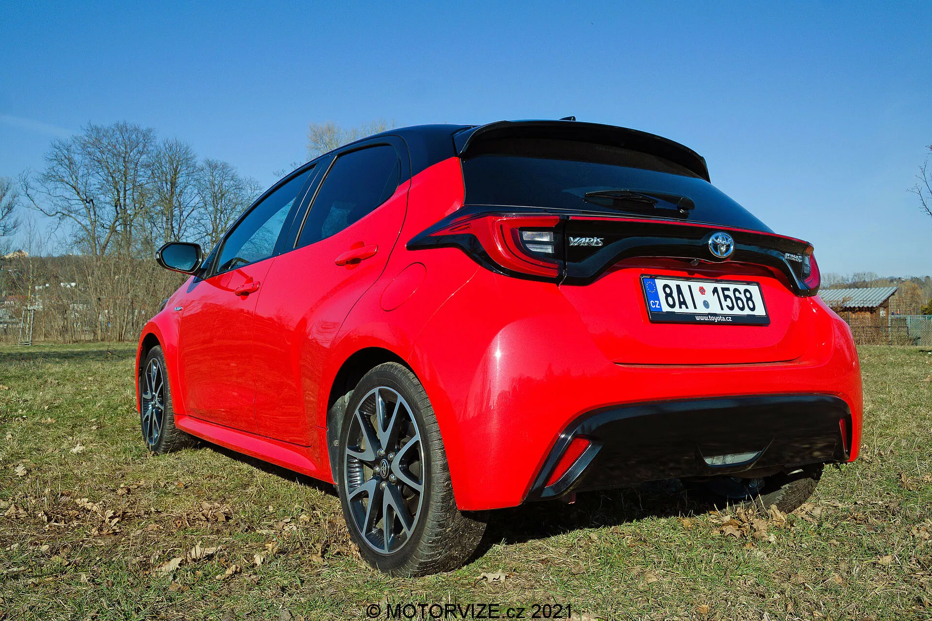 Zadnji tristranski pogled na svetlo rdeči hatchback Toyota Yaris 2020, gledano pod nizkim kotom, poudarjajoč oblikovne linije in ključne funkcije, kot so LED zadnje luči, zadnji spojler, antena v obliki plavuti morskega psa in temno obdelana večkraka platišča iz zlitine. Fotografija zajema podrobnost zadnjega dela in del stranskega profila v naravni dnevni svetlobi, poudarjajoč športno estetiko in sodobne elemente.
