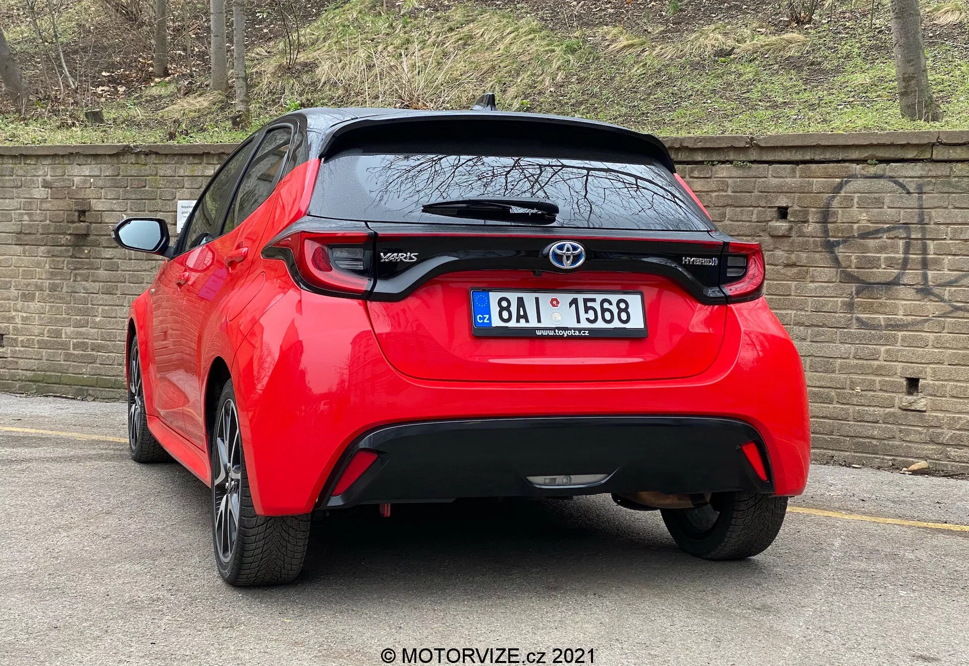 Visokoločljiv posnetek rdečega Toyotinega hatchbacka Yaris 2020, prikazanega iz zadnjega trikotnega kota. Vozilo ima značilne zadnje luči, osrednji Toyota emblem nad verjetno zadnjo kamero in oznako modela 'Yaris' na spodnjem levem delu vrat prtljažnika. Pod območjem registrske tablice je črni dekorativni kos, ki bi lahko bil iz plastike ali ogljikovega vlakna, majhen zadnji spojler pa se razteza nad zadnjim oknom. Avto, parkirano blizu roba na tlakovanem cestišču poleg opečnatega zidu, nakazuje urbano ali stanovanjsko okolje.