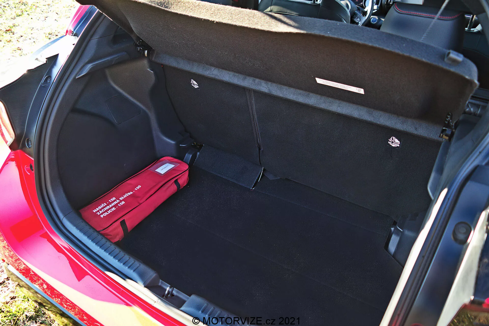 Prtljažnik rdečega Toyota Yaris 2020, viden iz višjega kota, ki prikazuje čisto, prostorno notranjost s črnim preprogo, odprtim pokrovom prtljažnika s potegovalnimi ročaji, ravno nakladalno talno površino, točkom za pritrjevanje tovora na desni in rdečim kompletom prve pomoči ali kompletom za nujne primere na levi. Ta dnevna fotografija poudarja veliko kapaciteto prostora in organizacijske funkcije vozila.