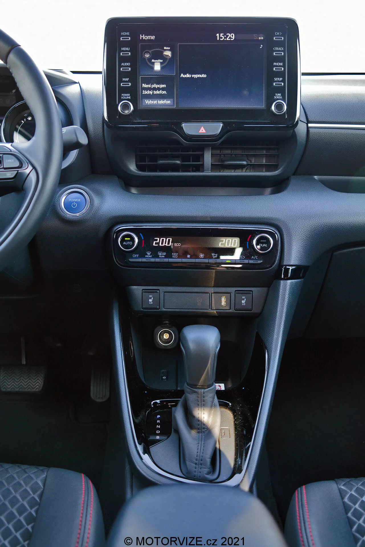 Pogled na notranjost Toyota Yaris 2020, osredotočen na centralno konzolo in elemente armaturne plošče. Fotografija, posneta iz prednjega sovoznikovega sedeža, prikazuje digitalni zaslon na dotik infotainment sistema, ki prikazuje domačo zaslonsko sliko s časom 15:29 in različnimi menijskimi možnostmi, centralne zračne reže in uporabniške kontrole za klimatizacijo in ogrevanje z digitalnim prikazom temperature nastavljeno na 20,0 stopinj. Centralna konzola vsebuje prestavno ročico oblečeno v usnje s srebrnim poudarkom in ročno zavoro. Notranjost je zasnovana v temnih tonih s kovinskimi zaključki, kar zagotavlja sodoben in prefinjen estetski videz.