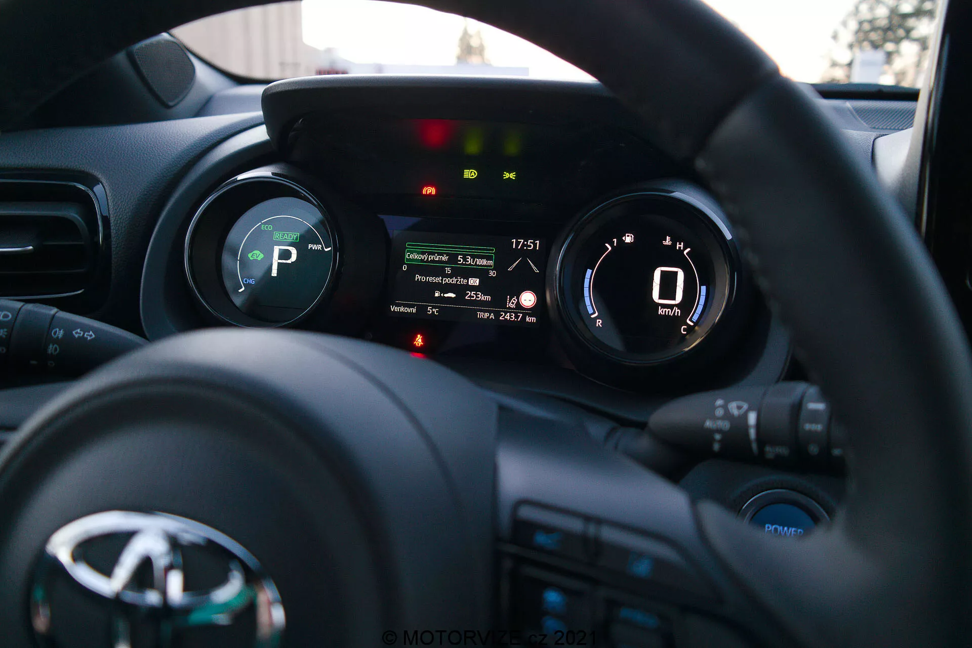 Fotografija notranjosti Toyote Yaris 2020 iz perspektive voznika, s poudarkom na volanu in armaturni plošči. Volan z logotipom proizvajalca in več gumbi za medije, komunikacijo in tempomat je spodaj. Armaturna plošča za volanom ima digitalni zaslon, ki prikazuje merilnik hitrosti s hitrostjo 0 km/h in indikatorje za prestavo (P za parkiranje), opomnik za varnostni pas, opozorilo o odprtih vratih in druge svetilke za stanje vozila. Majhna globinska ostrina poudari armaturno ploščo, medtem ko sta armaturna plošča in notranjost avtomobila rahlo zamegljeni v ozadju. Slika, verjetno posneta pozno popoldne ali zgodaj zvečer, prikazuje sodobno instrumentacijo in tehnologijo vmesnika vozila.