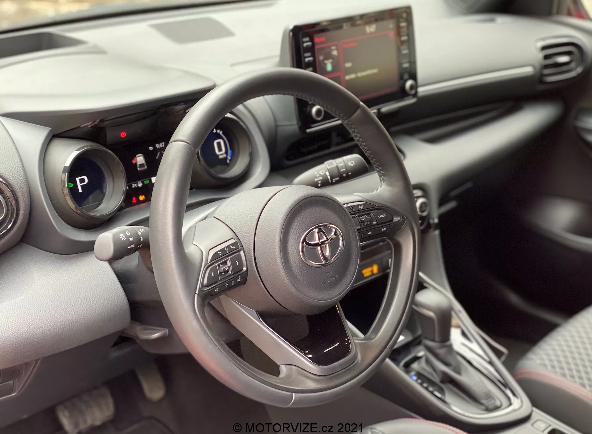 Pogled na notranjost Toyote Yaris 2020 z voznikovega sedeža, osredotočen na voznikov prostor. Slika poudarja trikraki volan Toyote z gumbi za upravljanje zvoka, Bluetooth in tempomata, s Toyotinim emblemom v sredini. Za volanom je delno viden digitalni instrumentni zaslon, ki kaže, da je avtomobil v načinu 'P' (parkiranje). Sredinska konzola prikazuje zaslon infotainment sistema in pod njim upravljalnike HVAC, skupaj z možnimi vhodi USB ali dodatnimi vhodi. Slika tudi prikazuje prestavno ročico avtomatskega menjalnika s sodobno zasnovo in usnjenim pokrovom. Notranjost ima sodoben dizajn s kombinacijo mehkih materialov in srebrnih poudarkov, skupaj z držali za pijače ali majhnimi odlagalnimi predali poleg prestavne ročice. Delno viden panel vrat razkriva nadzorno ploščo oken, kar nakazuje električne okna.