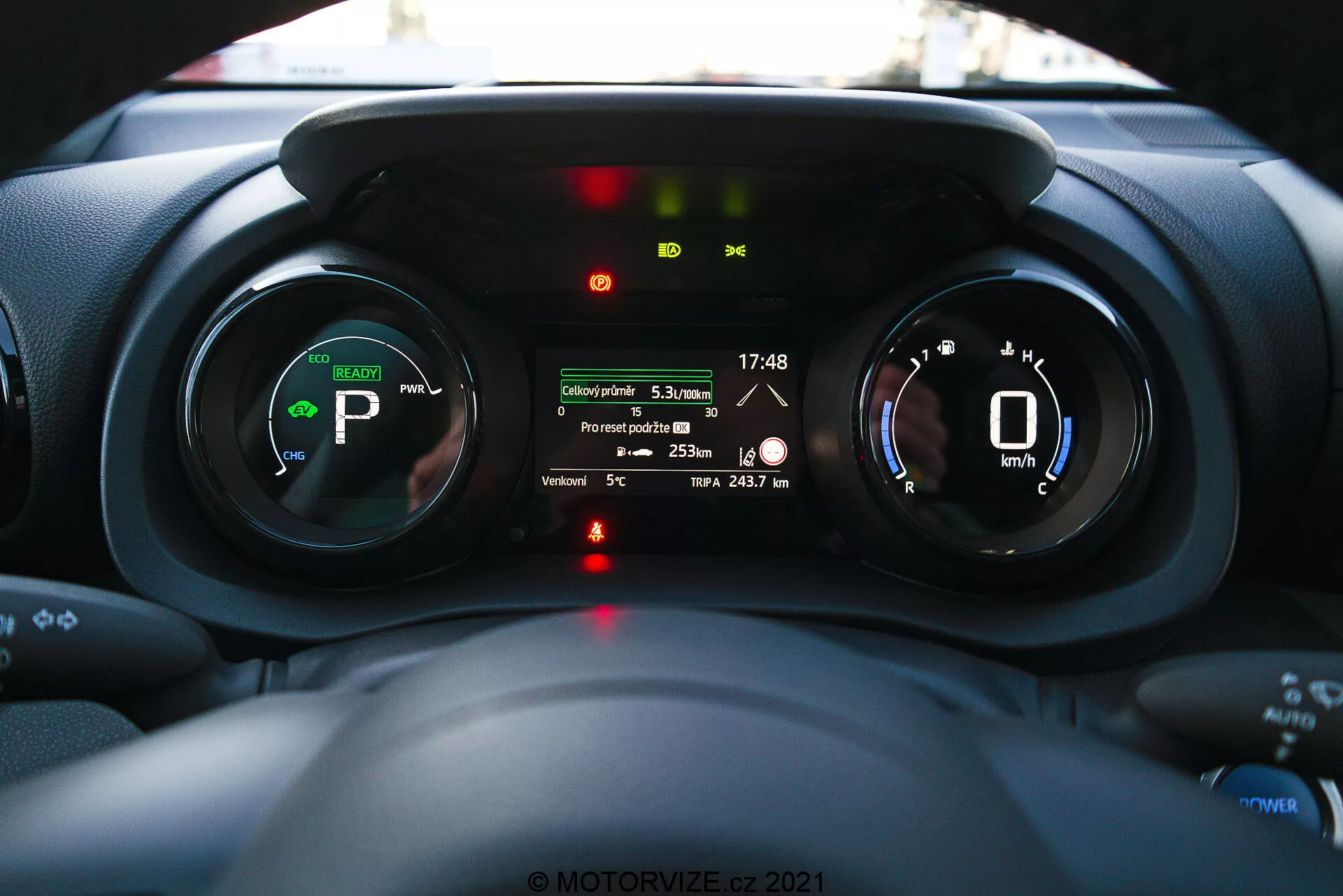 Notranji pogled na armaturno ploščo Toyota Yaris 2020 s poudarkom na instrumentni plošči, posnet iz perspektive voznika, prikazuje digitalne merilnike, indikacije za električno ali hibridno vozilo in večinformacijski zaslon s funkcijami, kot so razdalja poti, zunanja temperatura in doseg baterije.