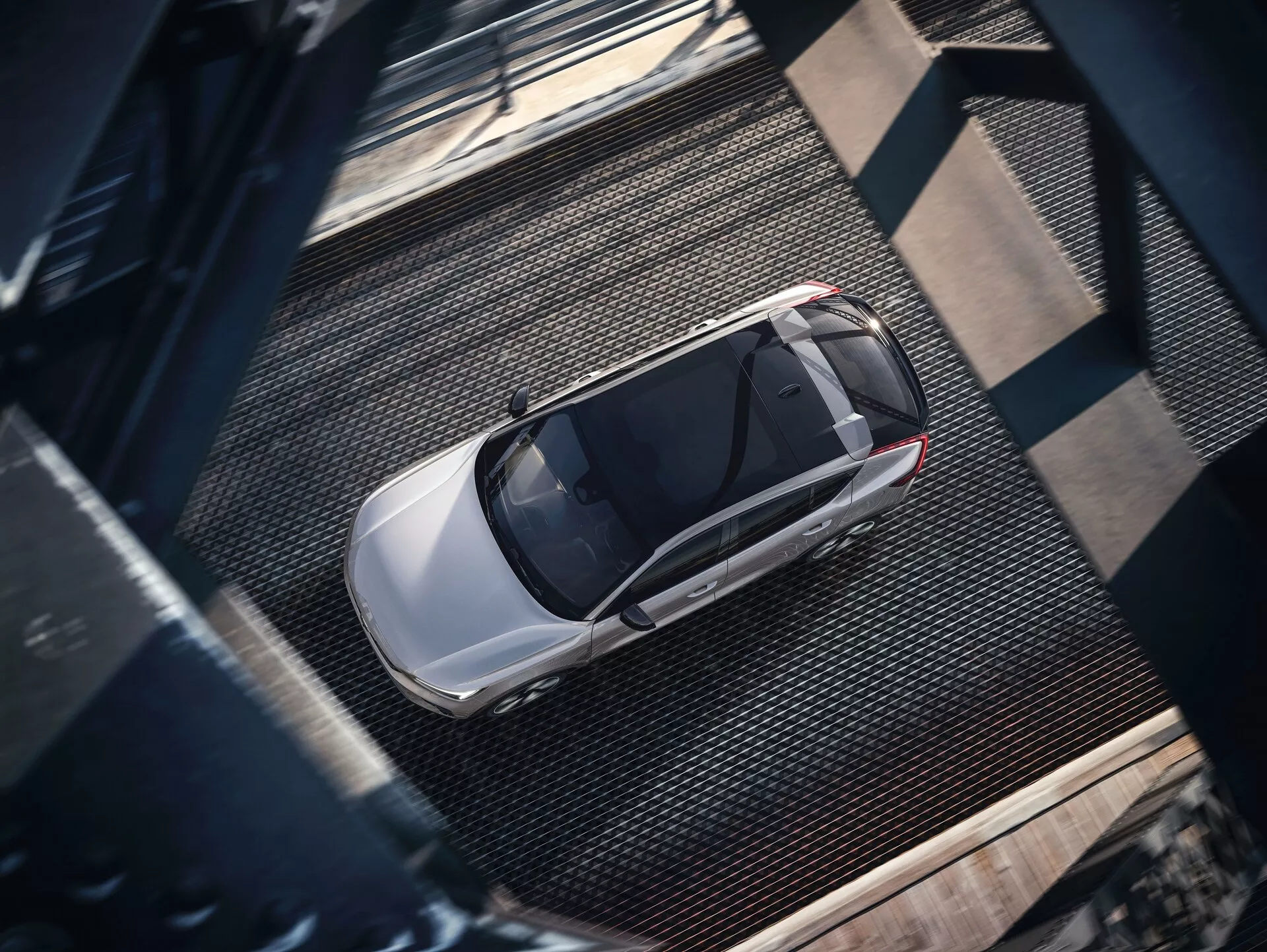 Pogled iz ptičje perspektive na Volvo C40 2021 prve generacije, ujet z visokega vidika. Slika razkriva celoten zgornji pogled na eleganten, sodoben limuzino, s svetlečo, kovinsko končno obdelavo, verjetno srebrno ali svetlo sivo barvo. Vidne lastnosti vključujejo streho, pokrov motorja, prtljažnik, okna in dele vrat, s prominentno streho, ki nakazuje napredno prezračevanje. Avto je parkiran na teksturirani industrijski površini z mrežastimi kovinskimi teksturami in temnimi zakovicami, ki mečejo sence, ki nakazujejo stransko osvetlitev, verjetno zjutraj ali pozno popoldne.
