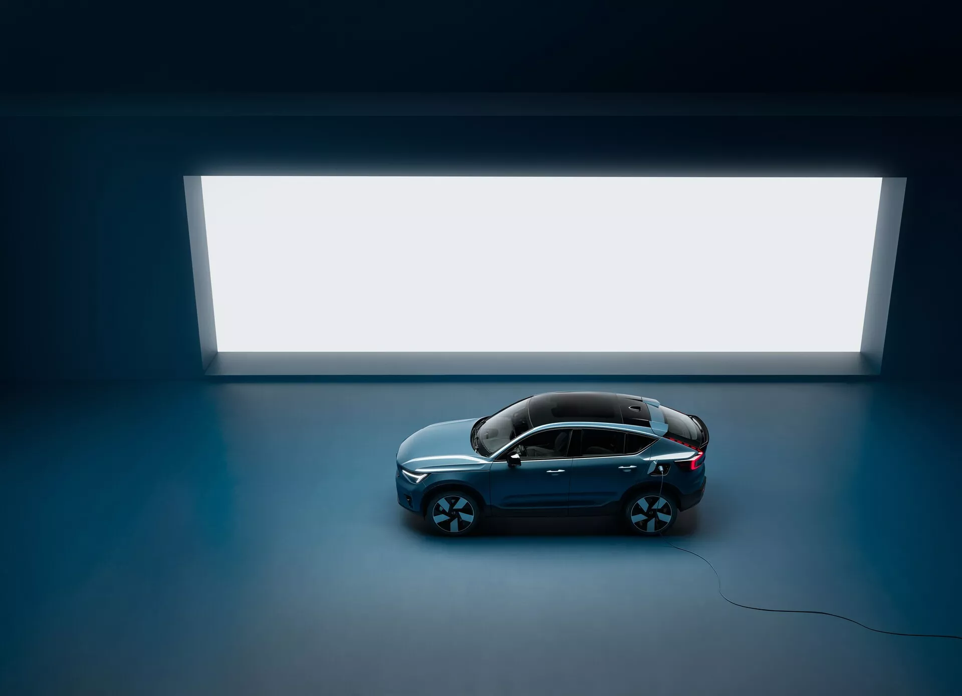 Posnetek iz visoke perspektive Volvo C40 2021 s štirimi vrati in panoramsko streho, posnet od zgoraj proti temni tleh z svetlim pravokotnim virom svetlobe v ozadju, ki prikazuje čist in minimalističen dizajn s temno, sijočo karoserijo ter velikimi večkrakimi platišči iz lahkih zlitin ter poudarjanjem njegovega razkošnega in sodobnega estetskega videza.