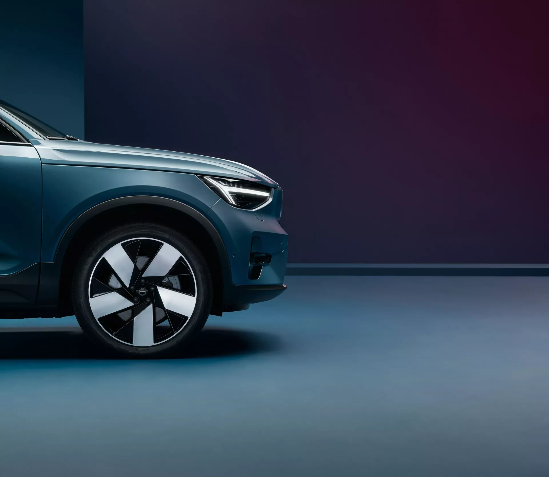 Nizkotonski posnetek Volvo C40 2021 (prva generacija), ki se osredotoča na prednjo četrtino od prednjega kolesa do sredine pokrova motorja. Poudarjeno je sodobno oblikovanje žarometov, večnaperni lita platišča, kovinski sijaj barve in aerodinamične linije. Dvobarvno ozadje z mehko osvetlitvijo poudarja premium končno obdelavo in močno držo vozila.
