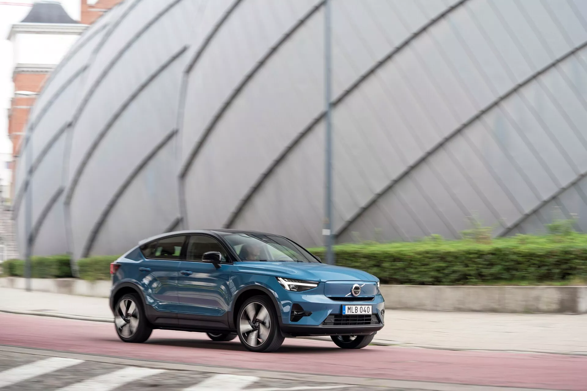 Slika Volvo C40 2021 (prva generacija) v gibanju po mestni cesti. Štirivratni crossover SUV je posnet iz kota treh četrtin spredaj, poudarjajoč elegantne, aerodinamične linije, oblikovane bočne panele in prepoznavna kolesa. Temno modra kovinska barva in izrazita prednja rešetka z emblemom proizvajalca nakazujeta možno električno ali hibridno različico. LED žarometi z ožkim, agresivnim dizajnom in vidnimi dnevnimi lučmi dodajo sodoben privlak. Ozadje z moderno arhitekturo poudarja urbano okolje.