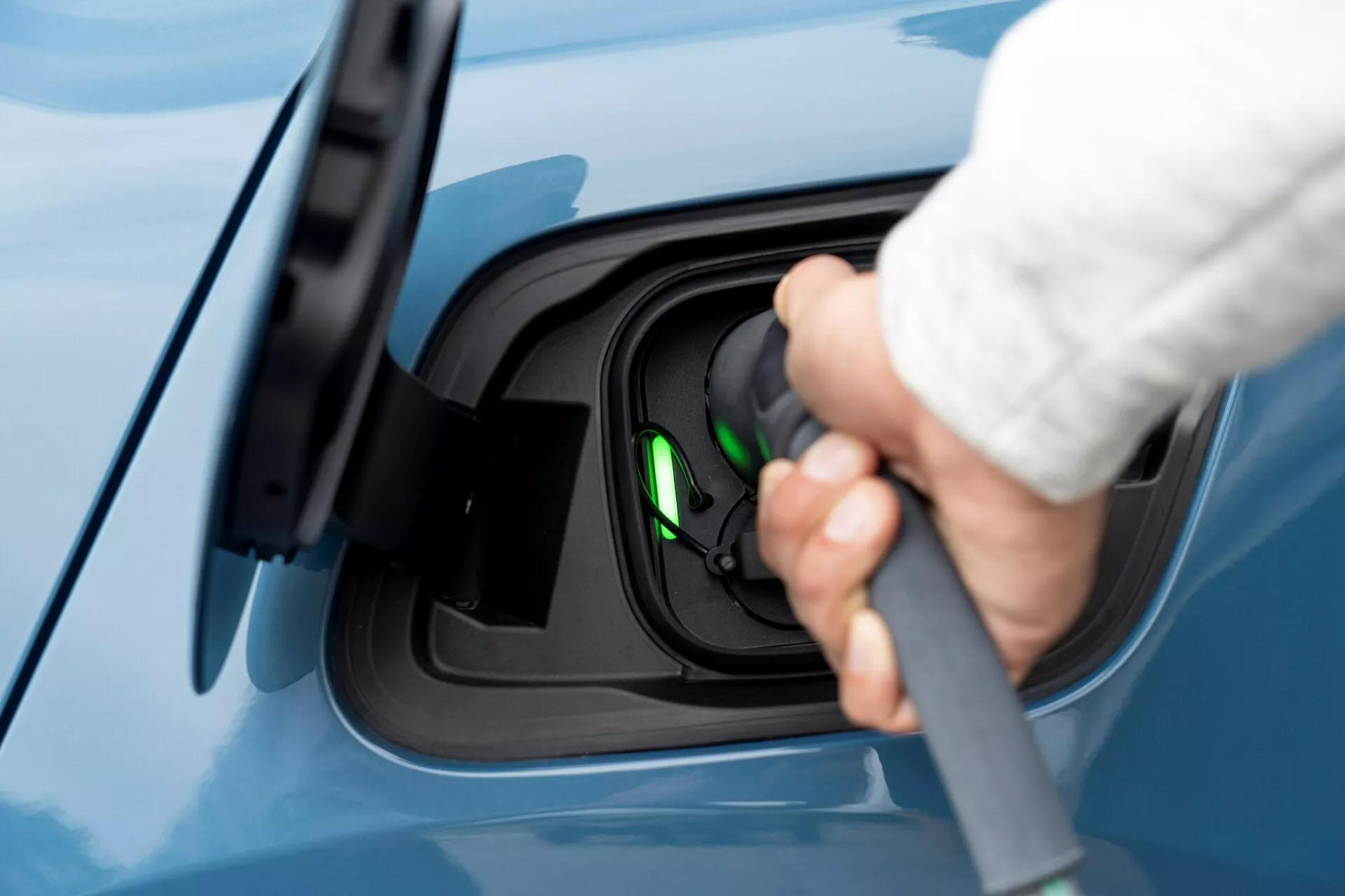 Bližinski stranski pogled na območje polnilnega priključka električnega vozila Volvo C40, letnik 2021 (prva generacija), z roko, ki priključuje polnilni kabel v priključek. Slika se osredotoča na polnilni proces, poudarja odprt polnilni priključek z verjetno uporabljanim tipa 2 spojnikom. Zelena lučka znotraj vtičnice nakazuje, da je vozilo pripravljeno za polnjenje ali se trenutno polni. Pogled je nekoliko dvignjen, prikazuje brezhiben modri lak vozila, kar poudarja čist in moderen videz električnega vozila.