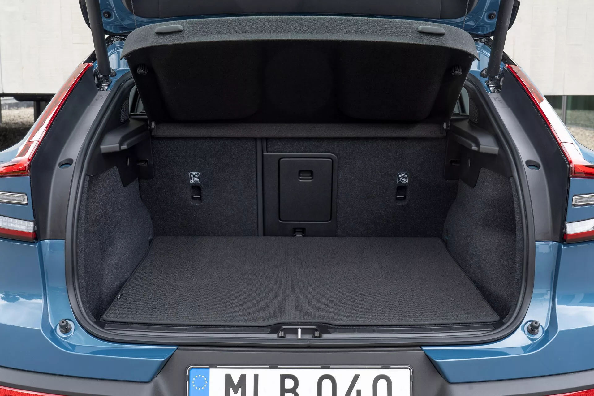 Prostor za prtljago sodobnega avtomobila, ki prikazuje prtljažnik Volvo C40 2021 (1. generacija) iz zadnje perspektive, rahlo pod nivojem pasu. Pokrov prtljažnika je odprt, poudarja standardno oblazinjenje notranjega obloka. Prtljažnik je prazen, prikazuje notranjost s preprogo, vdolbine ob straneh in ročaj na sredini tal za dostop do shranjevalnega prostora. Niso vidni nobeni kavlji ali pritrdilne točke za tovor. Evropska tablica na mestu predlaga evropski izvor.