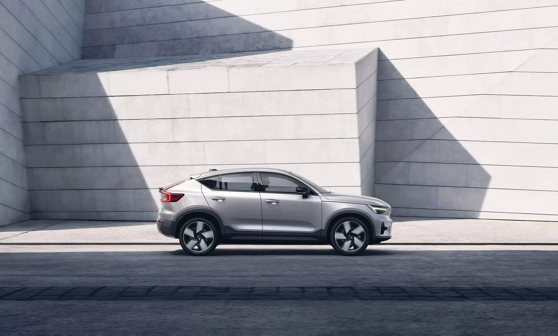 Slika modernega SUV Volvo C40 2021 parkiranega vzporedno, prikazuje levi stranski profil od levega sprednjega žarometa do leve zadnje luči. Kot poudarja odmik od tal in držo na asfaltni površini. Sončna svetloba ustvarja sence na sijajnem barvnem premazu. SUV ima eleganten dizajn z rahlo poševno linijo strehe, izrazitimi oblinami vrat, zatemnjenimi stekli in sodobnimi stranskimi ogledali. Platišča iz lahke litine z večkrakim dizajnom in pnevmatike z nizkim profilom kažejo na zmogljivost in luksuz. Vidni so bočni prag, obloge blatnikov in linija značaja, ki poteka skozi kljuke vrat, kar daje občutek robustnosti. Odsotnost vidnega izpuha nakazuje morebiten električni ali hibridni pogon. Minimalistične betonske strukture v ozadju kontrastirajo s prefinjenimi krivuljami avtomobila, kar poudarja njegovo urbano in oblikovno usmerjeno privlačnost.