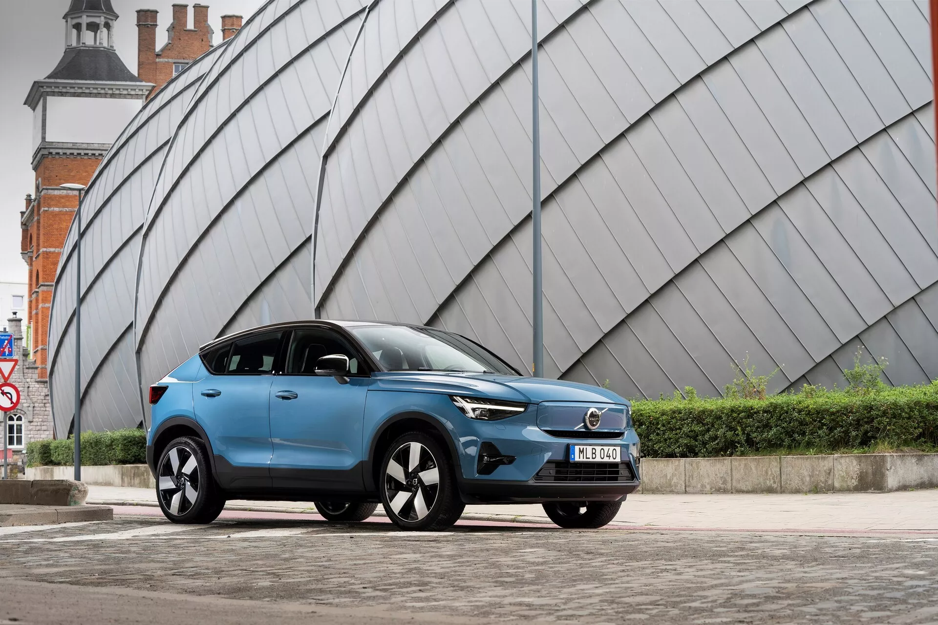 Moderno modro Volvo C40 2021 SUV parkirano na tlakovani mestni površini s kotno, kovinsko izgledajočo strukturo v ozadju. Nizek kot zajema s prednje leve strani prikazuje prednjo mrežo vozila, LED žaromete, aerodinamični profil, večkraka lita platišča, stranska ogledala z vgrajenimi smerniki, mat ali satenast zaključek barve, zatemnjena stekla, sprednjo registrsko tablico in izklesane linije bokov karoserije.