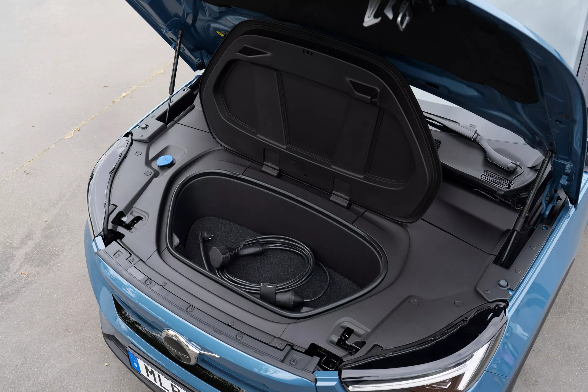 Bližnji, poševni pogled od zgoraj na Volvo C40 2021 (1. generacija) z odprtim frunkom, ki razkriva prazen shranjevalni prostor in zvito polnilno kabel, kar kaže, da gre za električno vozilo. Kovinska modra barva, notranjost pokrova frunka je črna z informativnimi nalepkami, delni pogled na logotip proizvajalca, elegantne žaromete in vidna sprednja rešetka, kar nakazuje na EV s sodobno LED tehnologijo in aerodinamično estetiko.