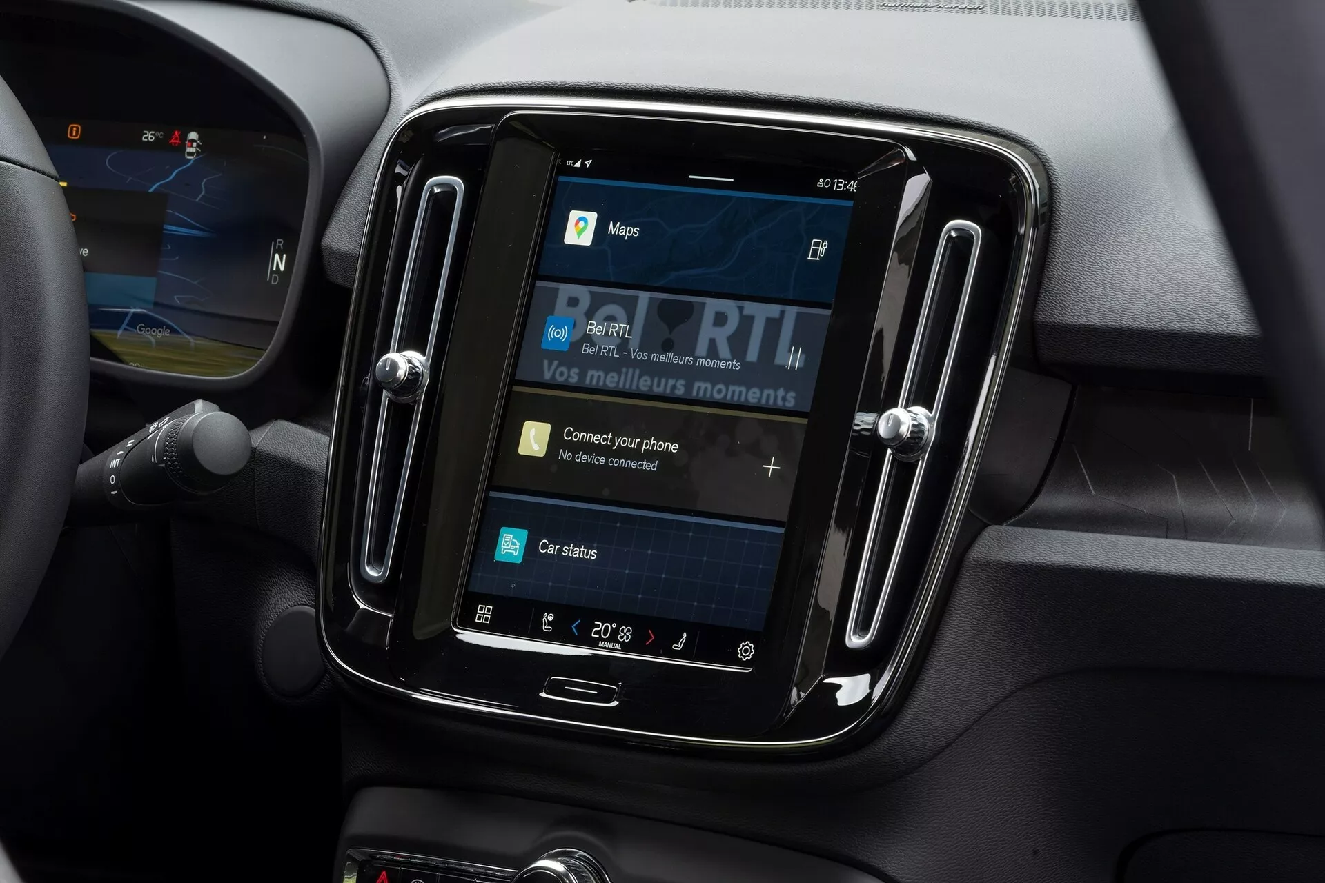 Pogled od blizu na informacijsko-razvedrilni sistem znotraj avtomobila Volvo C40 2021. Posnetek, narejen iz rahlega kotnega naklona navzdol, je osredotočen na zaslon na dotik, umeščen v svetlečem, črnem okvirju. Zaslon prikazuje možnosti, kot so zemljevidi, mediji, povezljivost telefona in stanje avtomobila, s 'Poveži svoj telefon' kot interaktivnim elementom. Okoli informacijsko-razvedrilnega sistema so materiali z mehkim dotikom in obrisne linije, ki kažejo na prezračevalne odprtine, kar nakazuje na sodoben notranji del avtomobila. Nastavitev temperature kaže 20° Celzija, na levi strani pa se vidi del volana.