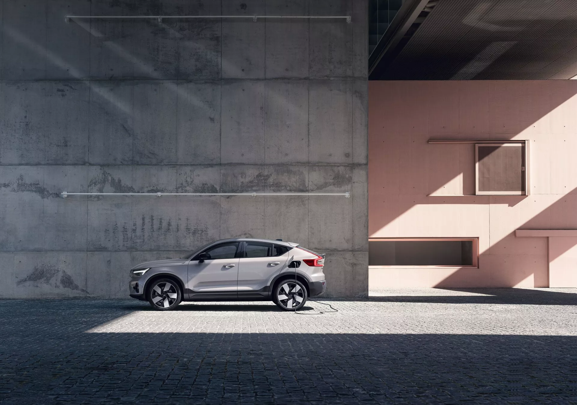 Stranski profil elegantnega SUV-ja Volvo C40 iz leta 2021 (1. generacija) proti urbanemu ozadju z betonskimi in breskovi zidovi. Avto ima odsevni bel premaz, elegantna lita platišča, zatemnjena okna, srebrne strešne nosilce in vgrajene smerne kazalce v vzvratnih ogledalih. Fotografija poudarja oblikovalske linije in krivulje avtomobila pod naravno dnevno svetlobo, kar dodaja občutek razkošja in prefinjenosti.