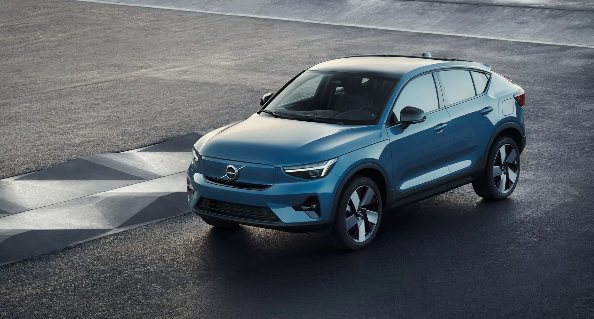 Pogled iz visokega kota na sodoben SUV Volvo C40 2021 (1. generacija) parkiran na asfaltni površini, ki prikazuje visoko širok sprednji pogled. Elegantno prednje območje, verjetno značilno za električna vozila z minimalnimi vstopi zraka, združuje ostre LED žaromete, gladko poševno pokrovo motorja in profilirane stranske linije z velikimi aluminijastimi platišči. Stranska ogledala z vgrajenimi smerniki, obilje steklenih površin z možno zatemnitvijo stekel in nežno nagnjena strešna linija s strešnimi nosilci so opazne lastnosti. Jasni pogled zajame luksuzno oblikovanje vozila, napredno aerodinamiko in sodobne tehnološke elemente.