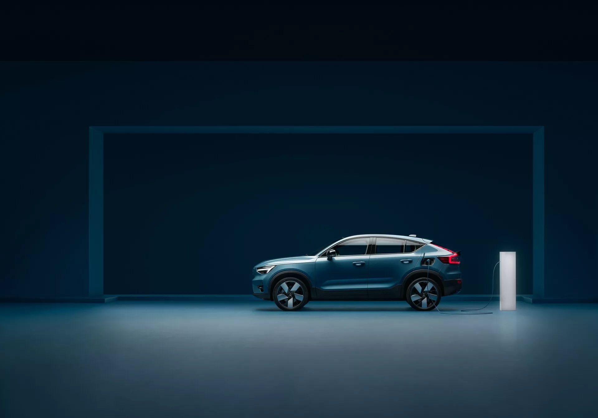 Pogled s strani na sodoben avtomobil Volvo C40 2021 (prva generacija) v studijskem okolju z odsevno talno površino, ki prikazuje eleganten dizajn, kovinsko barvo temno modre barve, poševno strešno linijo, velika večkraka aluminijasta platišča, oblikovane linije, robustno zgradbo, integrirana stranska ogledala s smerokazi, LED zadnje luči in polnilni priključek za elektriko.
