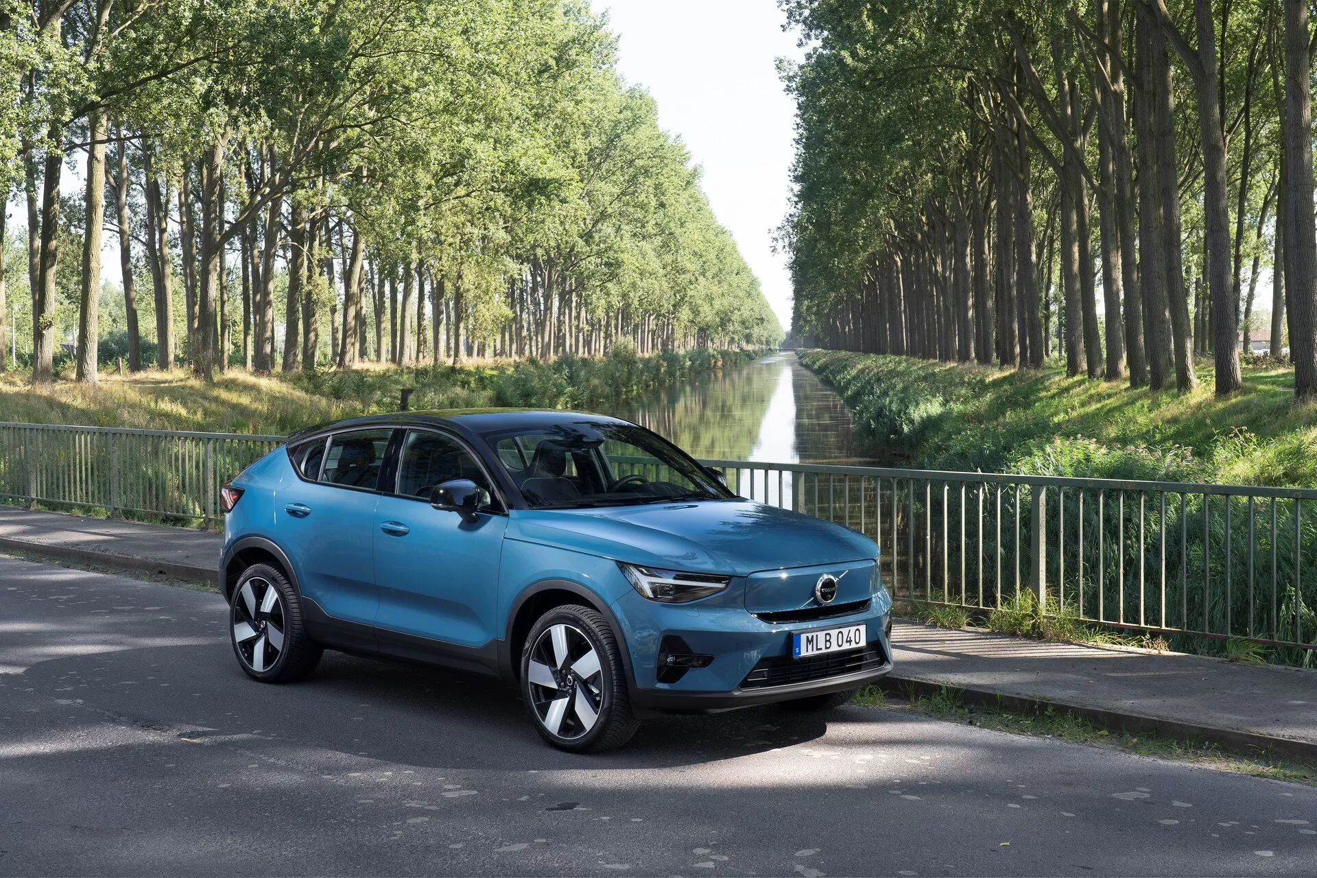 Volvo C40 2021 (Prva Generacija) kompaktni SUV v tričetrtinskem pogledu od spredaj levo na asfaltni cesti. Predstavlja sodobne oblikovalske elemente, kot so izrazita maska z osrednjim znakom, elegantne LED žaromete, stilska aluminijasta platišča, bočna ogledala z vgrajenimi smerokazi, aerodinamično oblikovana karoserija s svetlečo modro barvo, zatemnjena okna in štiri vrata. Osvetljeno v zunanjem okolju s cestnimi drevesi, osredotočeno na estetske in tehnološke lastnosti vozila.