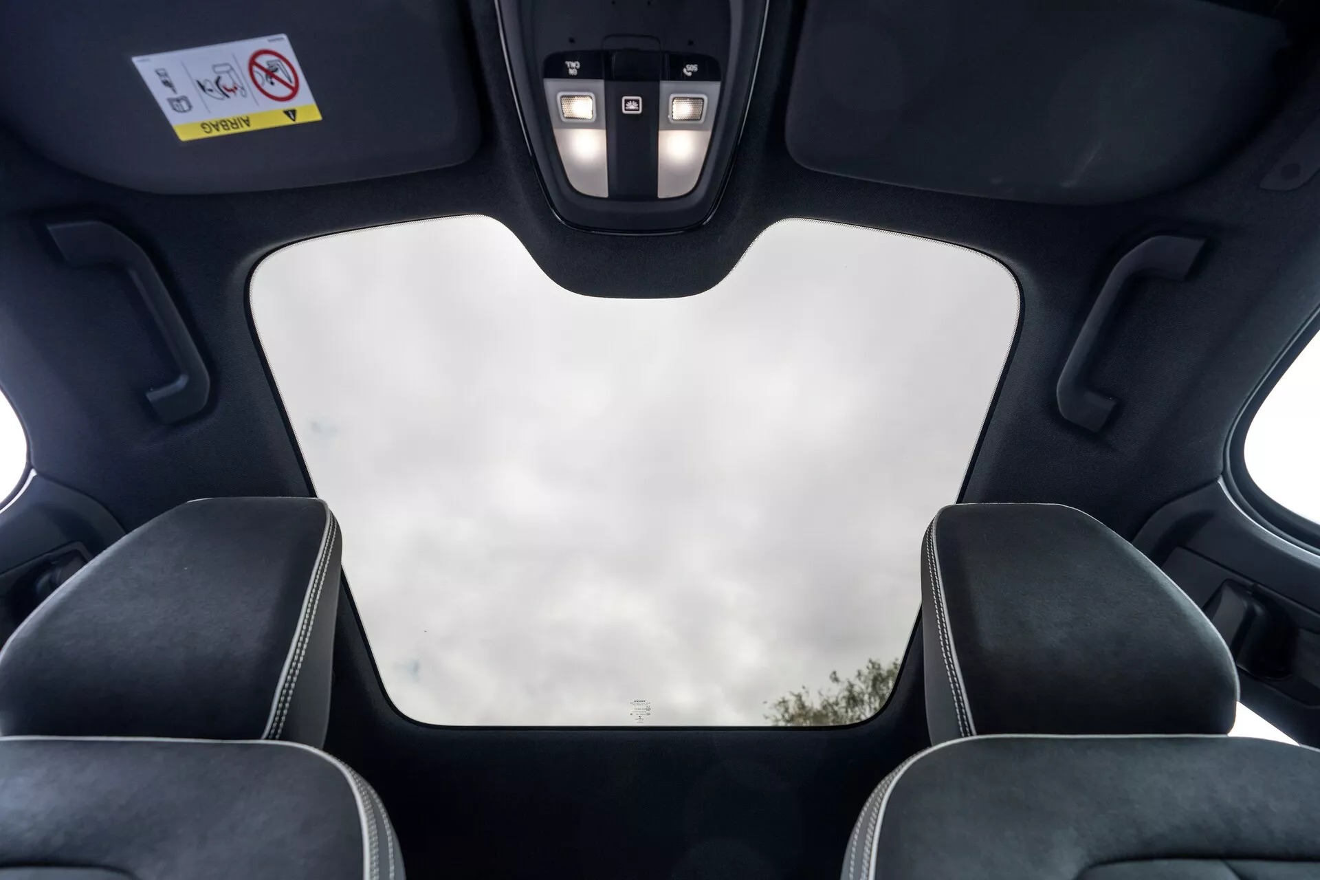 Notranji pogled na Volvo C40 2021 (1. generacija) iz perspektive zadnjega potnika, ki prikazuje strešne značilnosti, kot so zaprta panoramska streha z jasnim pogledom na nebo, črna obloga, izrezi za strešni mehanizem, LED osvetlitev in nadzorna plošča z gumbi za različne funkcije. Slika izpostavlja tudi vzglavnike sprednjih sedežev s kontrastnimi šivi, poudarjajoč moderni luksuzni dizajn vozila in njegove vrhunske lastnosti.