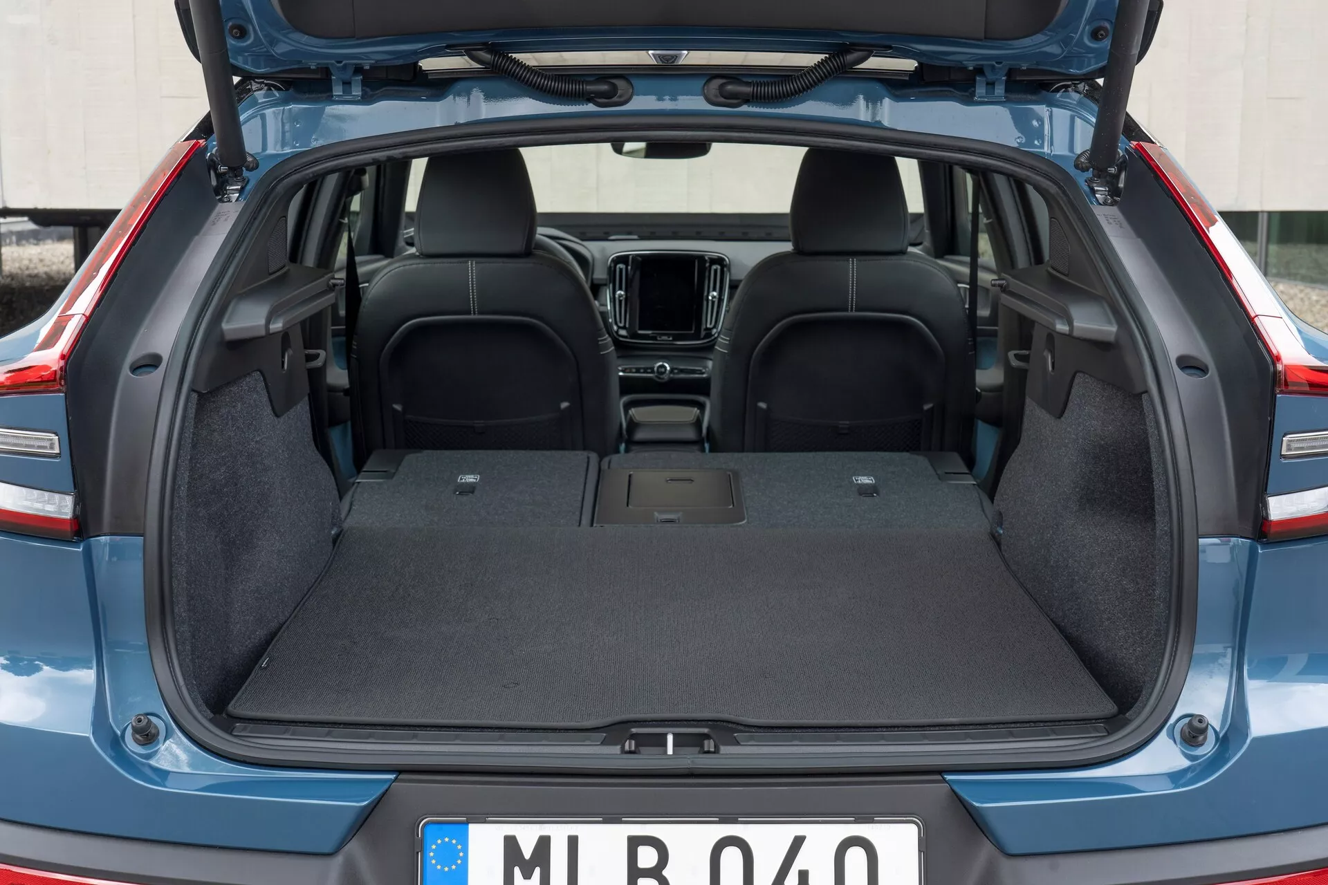 Zadnji notranji del Volvo C40 2021 (1. generacija) z odprtim prtljažnikom, ki prikazuje prazno prtljažno območje z vzdržljivo preprogo, ravno površino in brez vidnih vdorov. Videti je zgornji del zadnjih sedežev iz temne tkanine, s parcialnim pogledom na elemente prednje kabine, kot sta sredinska konzola in info-zabavni sistem. Pokrov prtljažnika je dvignjen, kar razkriva robove iz plastike/kompozita.