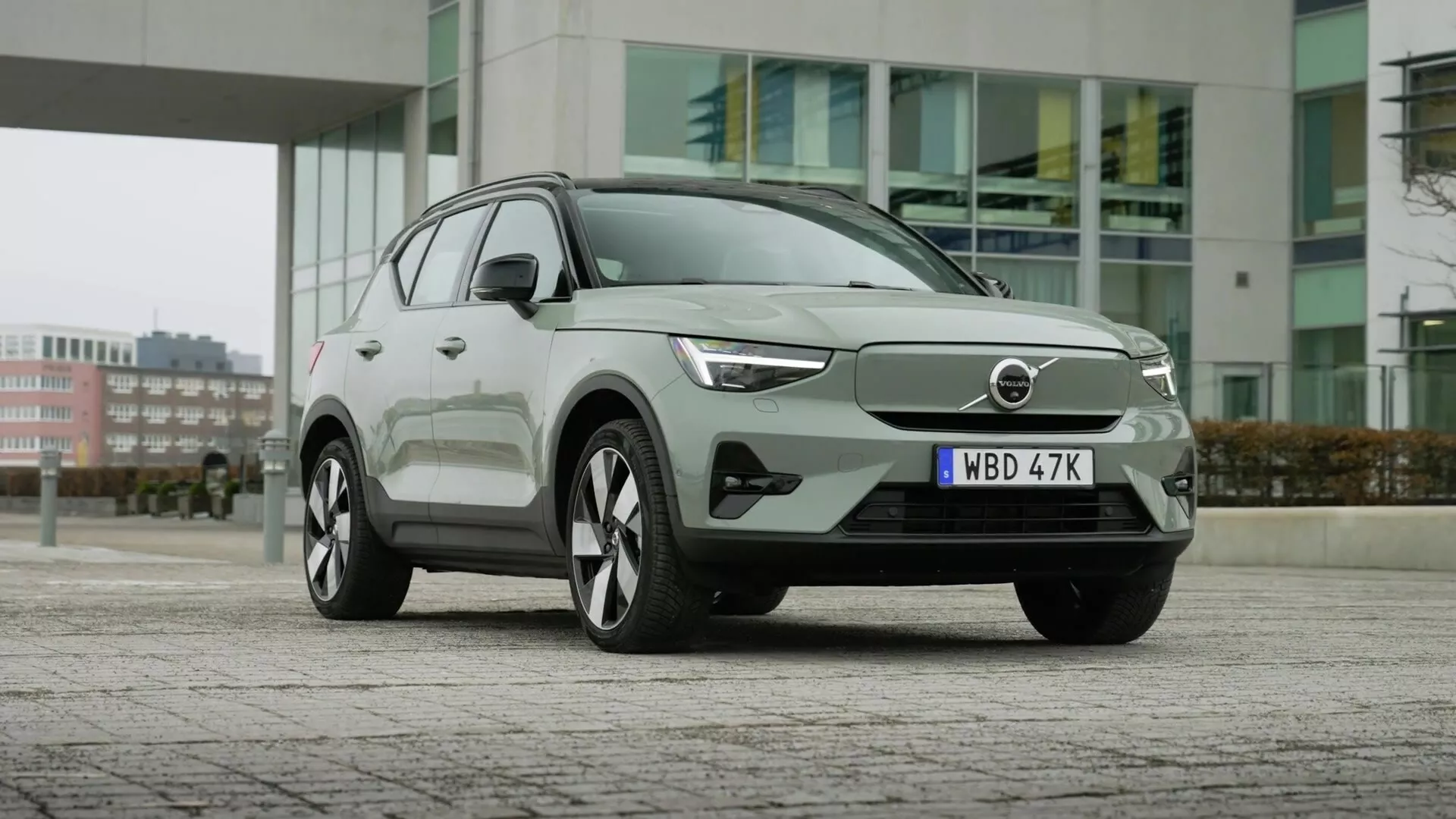 Električni SUV Volvo EX40 1. generacije letnik 2024, parkiran na tlakovcih z mestnimi stavbami v ozadju, prikazuje aerodinamično sprednjo fasado z zaprto rešetko, elegantne LED žaromete, velika večkraka lita kolesa, moderno obliko karoserije z ročaji vrat v ravni s telesom, integrirane smerne utripalke v stranskih ogledalih, okna z okvirji iz kroma in napredno tehnologijo pomoči vozniku.