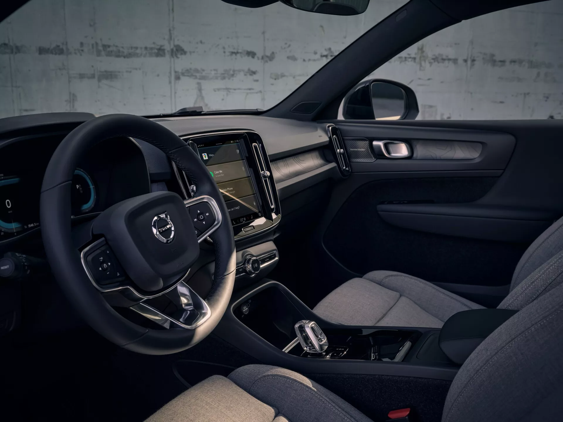 Pogled na notranjost Volvo EX40 prve generacije za leto 2024 z volanom na levi strani, digitalno instrumentno ploščo, navpičnim zaslonom na dotik za informacijsko-razvedrilni sistem, minimalističnim dizajnom armaturne plošče, udobnimi sedeži z bočno oporo, avtomatskim menjalnikom z ročnim načinom prestavljanja, vgrajeno ambientalno razsvetljavo, barvno paleto v temnih tonih in naprednimi tehnološkimi elementi.
