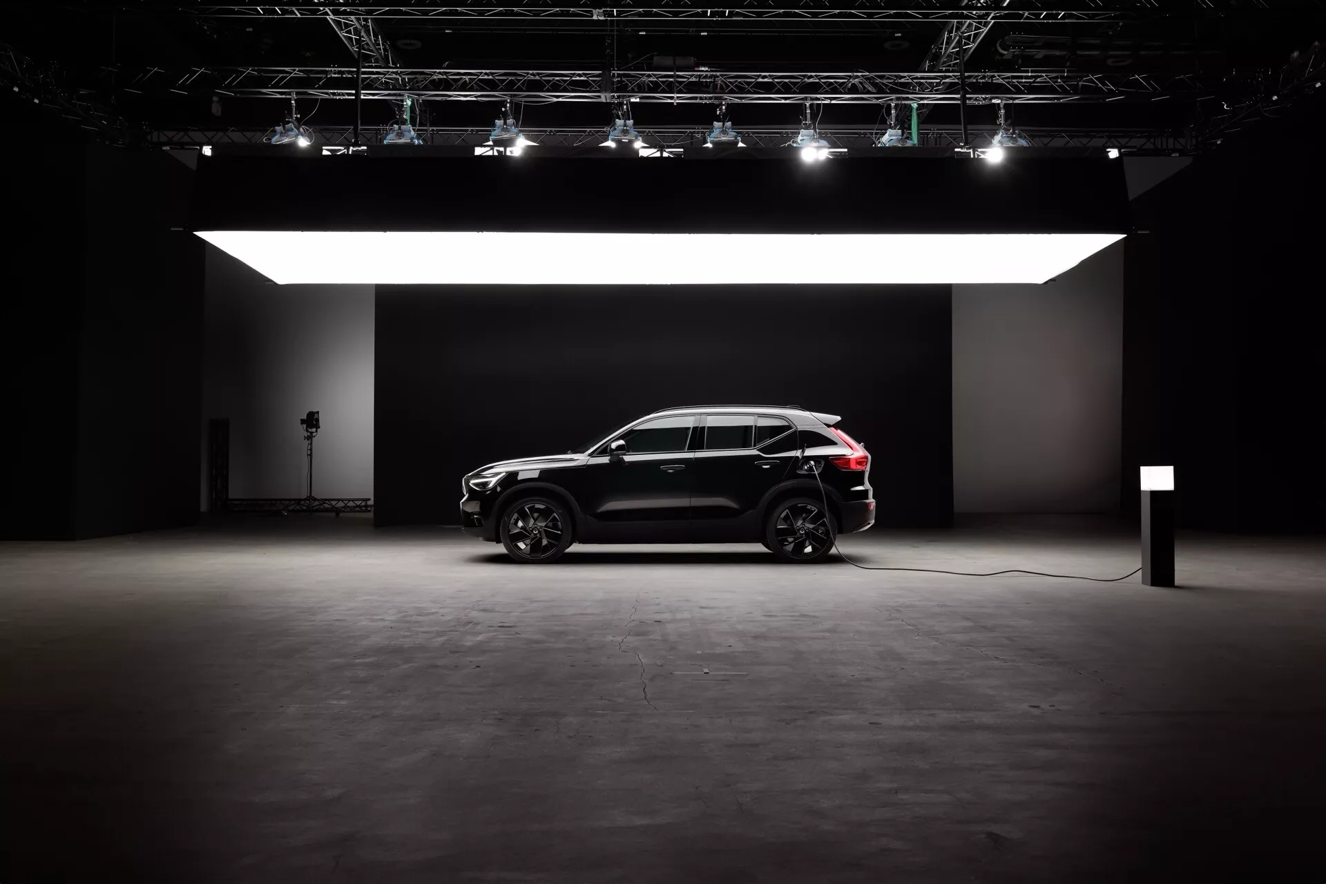 Stranski profil Volvo EX40 1. generacije 2024 v studijskem okolju, prikazuje njegovo elegantno črno zunanjost, sodobno oblikovanje SUV z športnim križanskim stilom in električne/hibridne zmogljivosti. Značilnosti vključujejo velika kolesa z večkrakim zlitinskim dizajnom, dramatično osvetlitev brez bleščanja in polnilni kabel, ki nakazuje tehnologijo trajnostne mobilnosti.