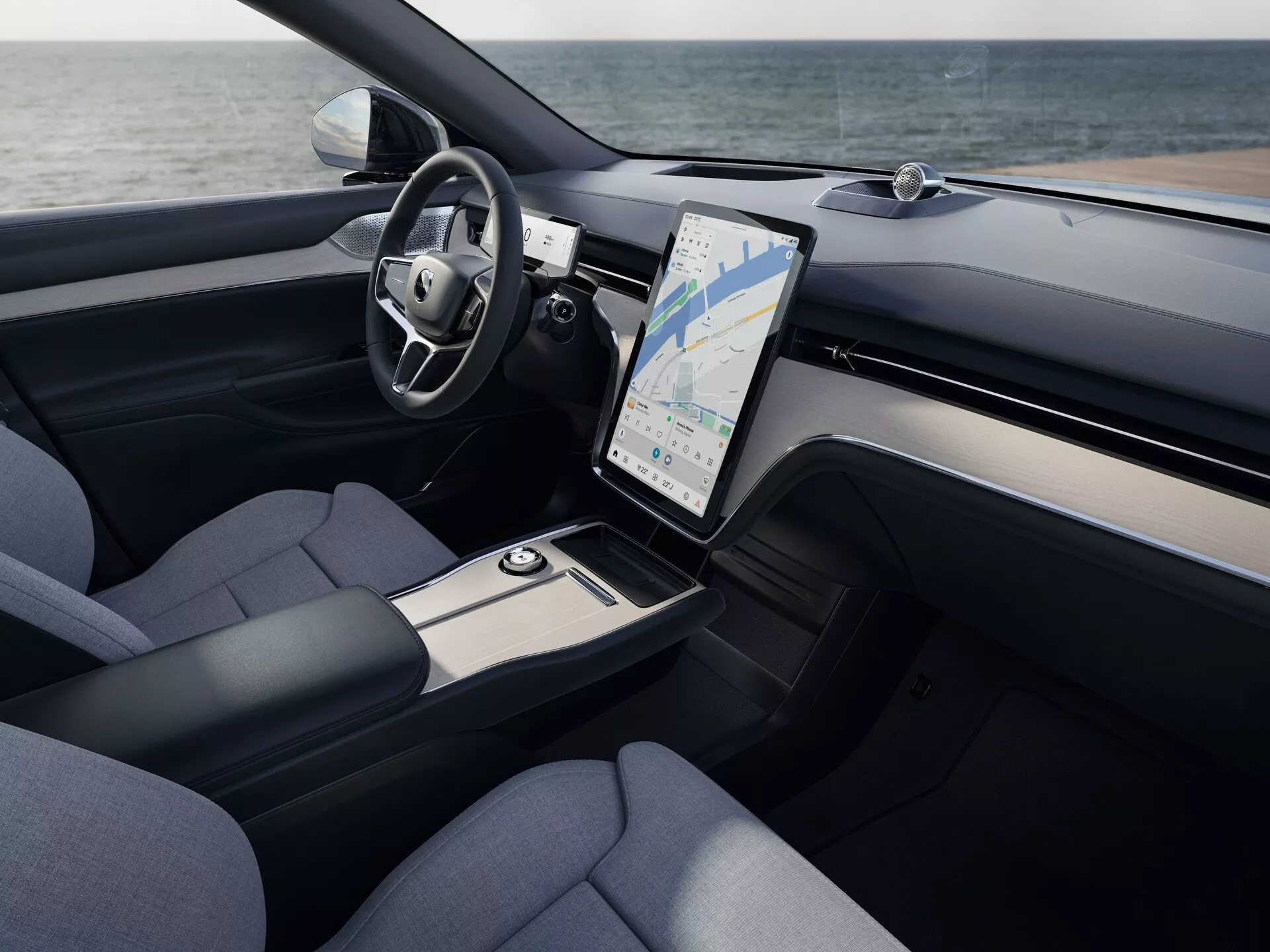 Pogled na notranjost Volvo EX90 2023, ki prikazuje območje voznika in sredinsko konzolo z višje perspektive, vključno z multifunkcijskim usnjenim volanom, velikim navpično usmerjenim zaslonom na dotik za informacijsko-razvedrilni sistem z GPS, prezračevalnimi rešetkami za klimo, poudarki iz brušenega kovina, sedeži oblazinjenimi z blagom in elegantnimi rešitvami za shranjevanje.