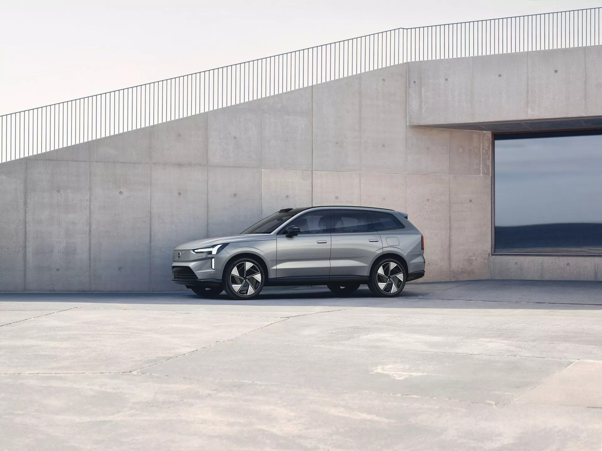 Volvo EX90 1. generacije letnik 2023, parkiran na prostem, stranski profil prikazuje kotni dizajn, ročaje vrat v isti ravnini kot karoserija in aluminijasta platišča ob minimalistični arhitekturni kulisi